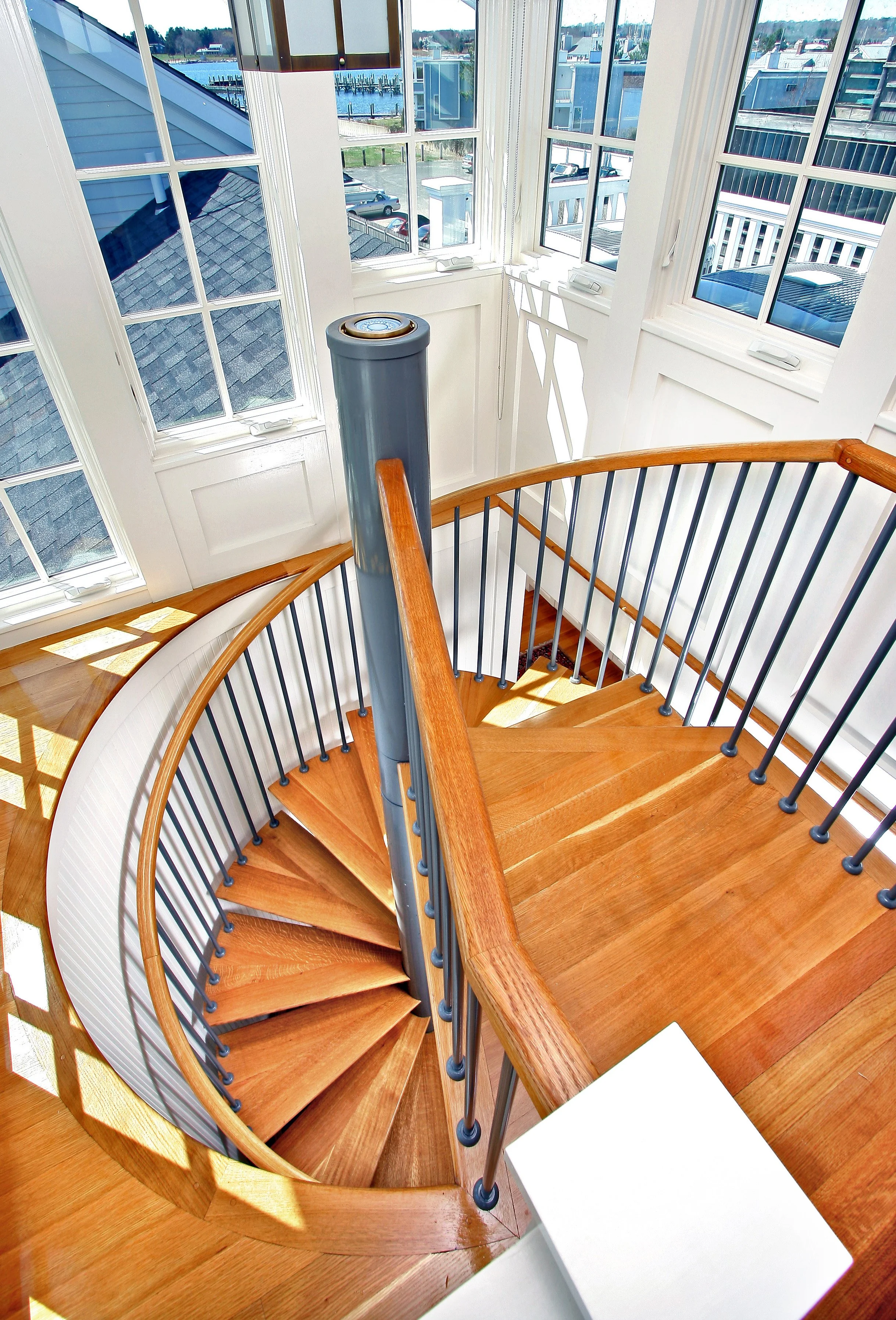 A spiral staircase made of wood with a wooden handrail and metal balusters, located near large windows with a view of a harbor with boats and buildings outside.