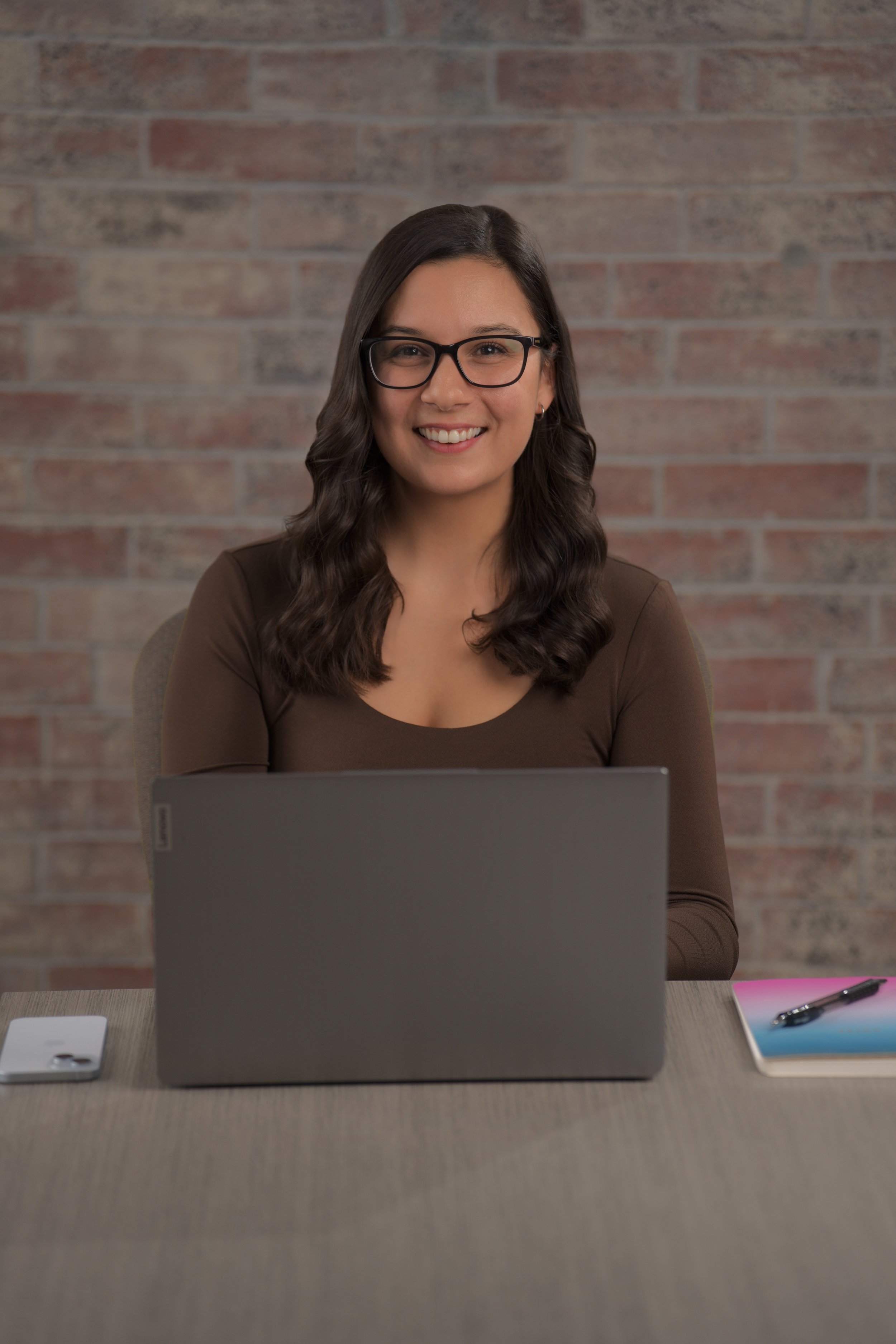 A woman with long dark wavy hair and glasses sitting at a desk in front of a laptop, smiling, with a notebook and pen on her right and a smartphone on her left, against a brick wall background.