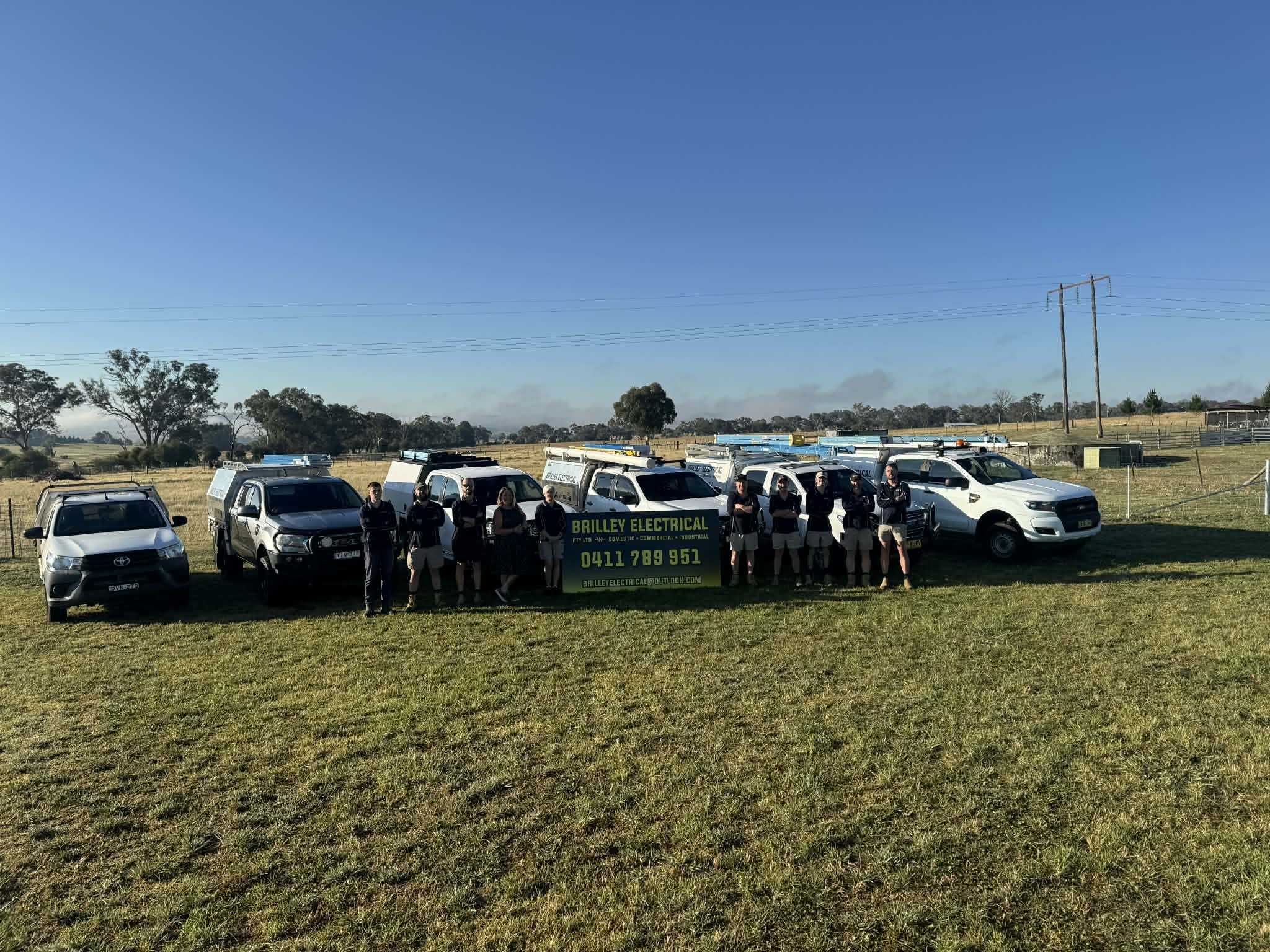The Brilley Electrical team standing in front of their fleet of service vehicles outdoors with a sign that reads 'BRILLEY ELECTRICAL' and a phone number, on a grassy field with a blue sky and trees in the background.