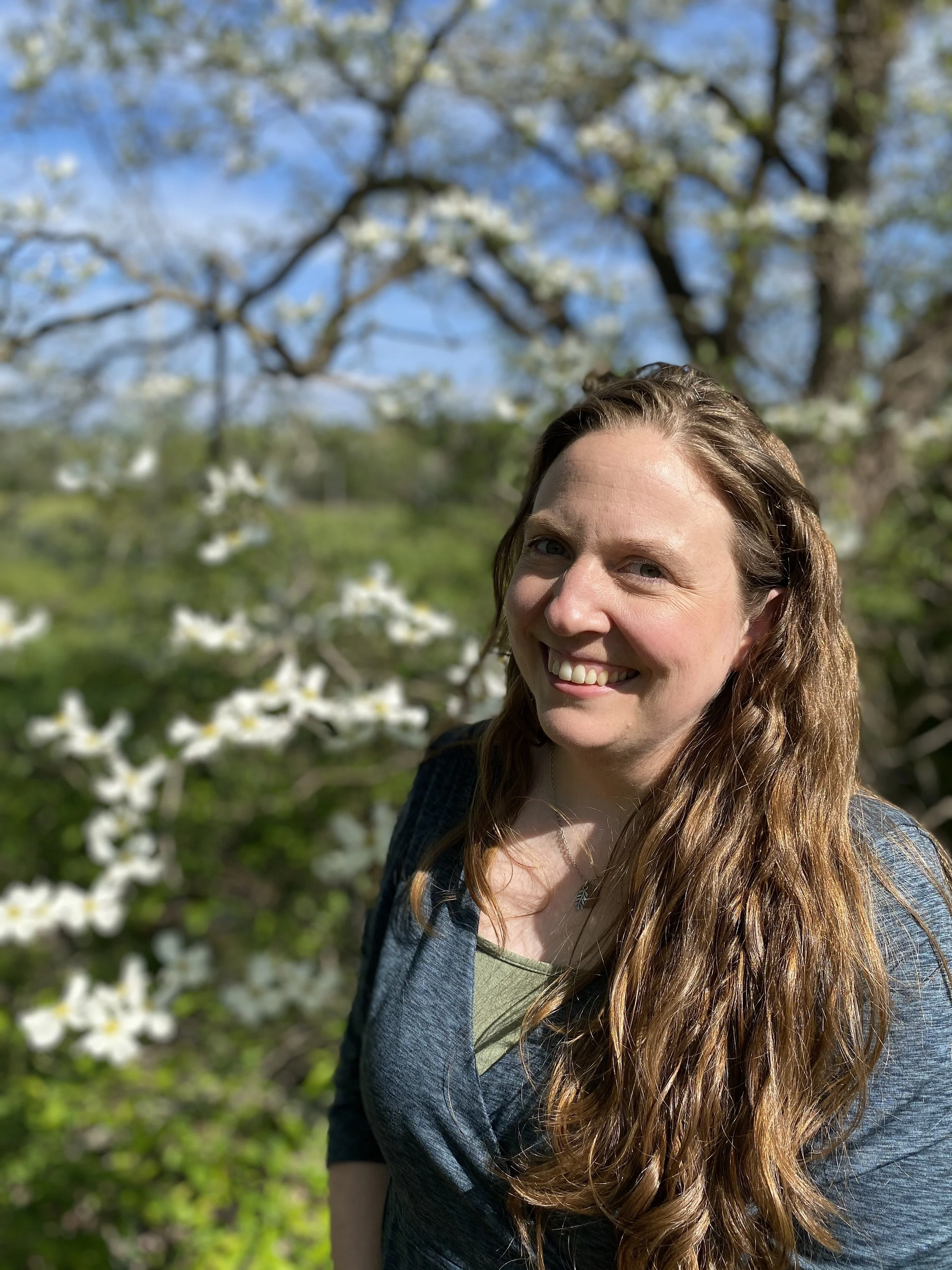 A woman with long brown hair smiling outdoors with blooming trees and a blue sky in the background.