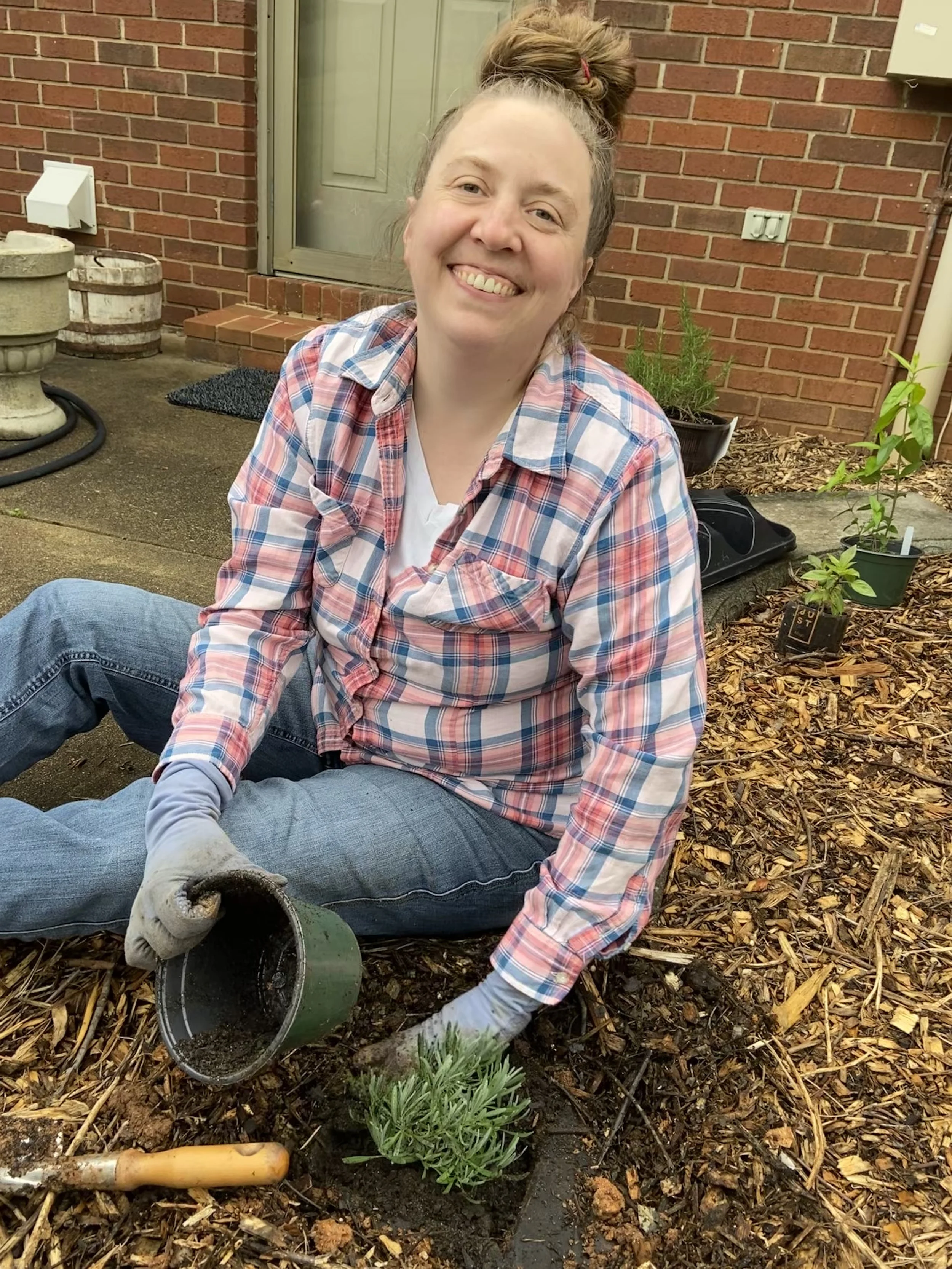 A woman smiling and planting a small plant in a garden bed, wearing gardening gloves, a plaid shirt, and jeans.