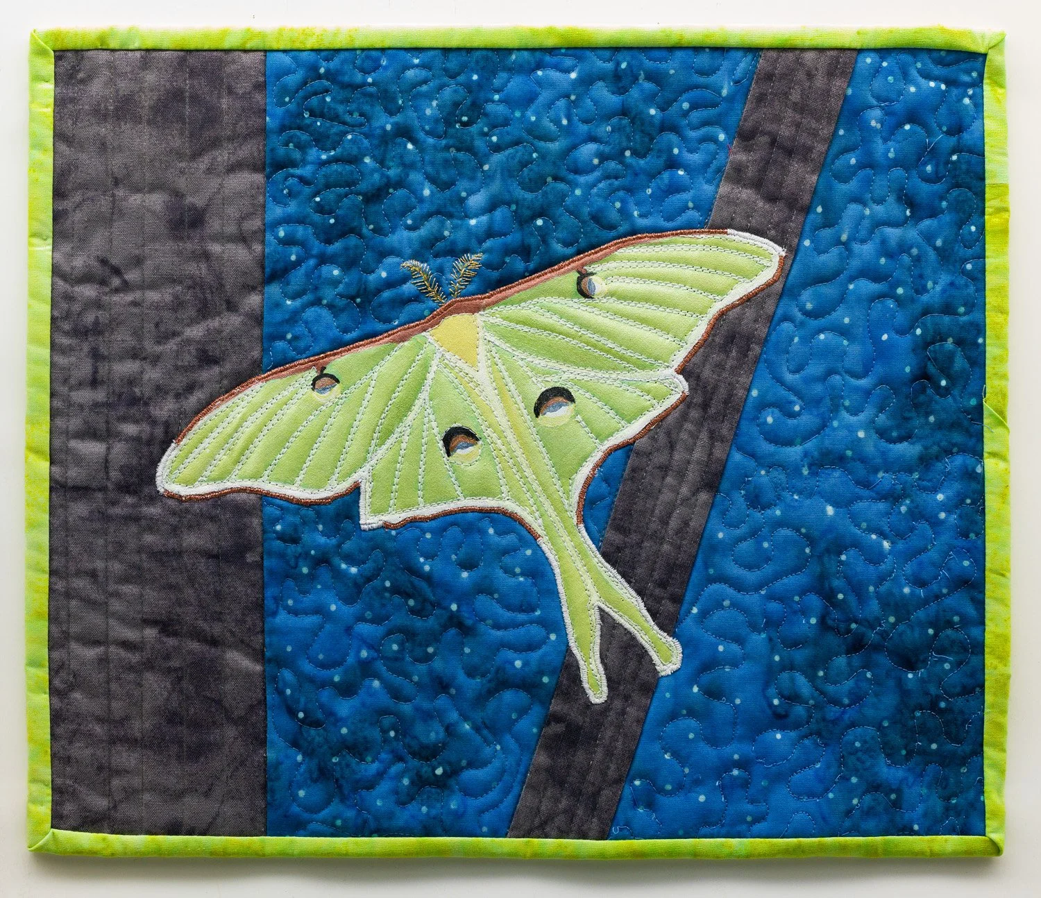 A quilted wall hanging featuring a large, green Luna moth with black and white eyespots on its wings, set against a background of blue and black fabric with a quilted swirling pattern.