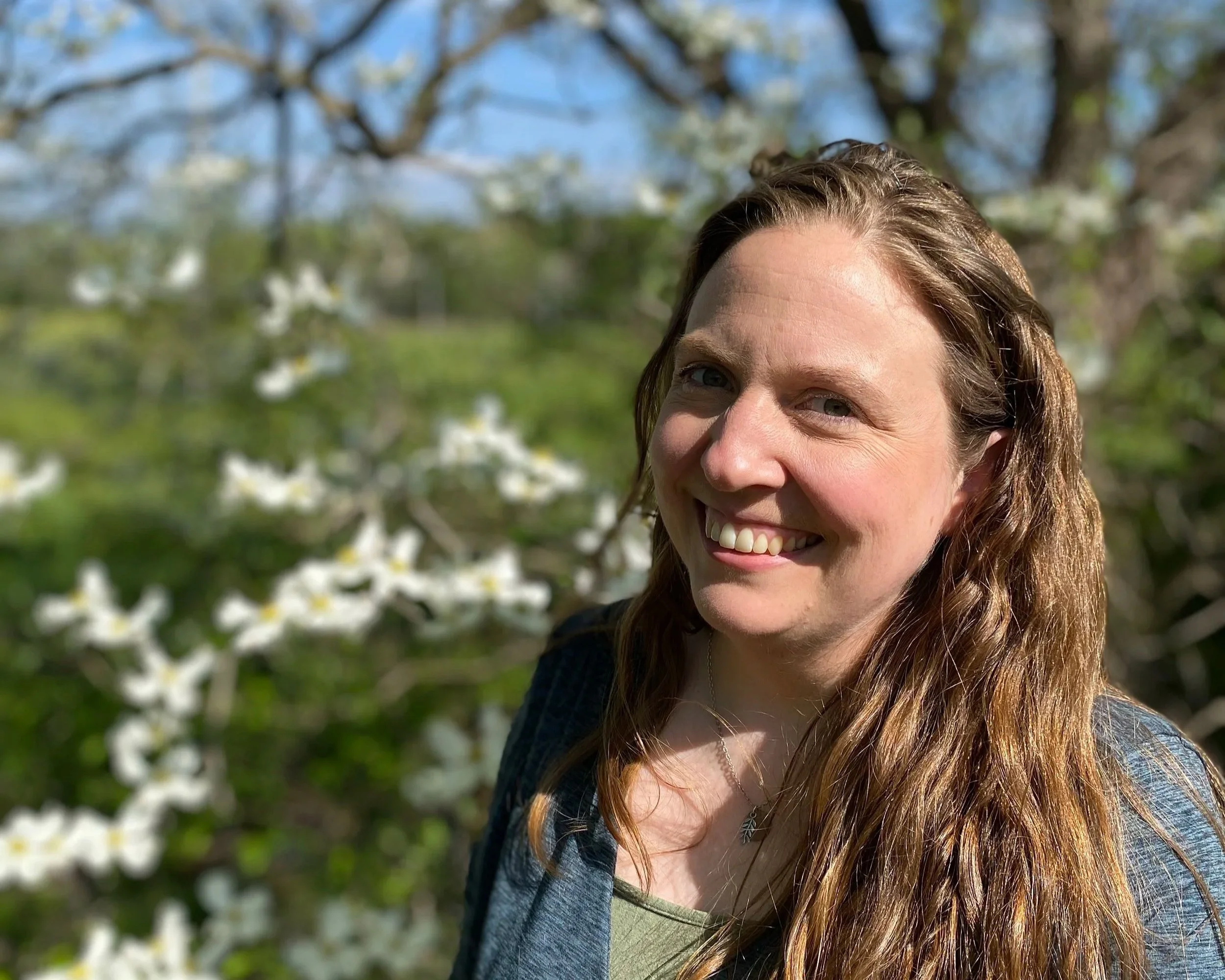 A woman with long wavy red hair smiling outdoors with white flowering trees and green foliage in the background.