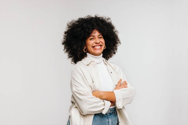 Smiling woman with curly hair wearing a white turtleneck, light jacket, and jeans, standing against a plain background.