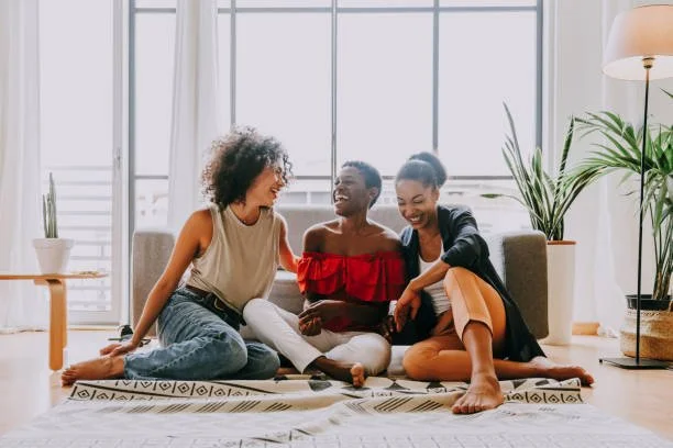 Three women sitting on a rug in a bright living room, smiling and enjoying each other's company.