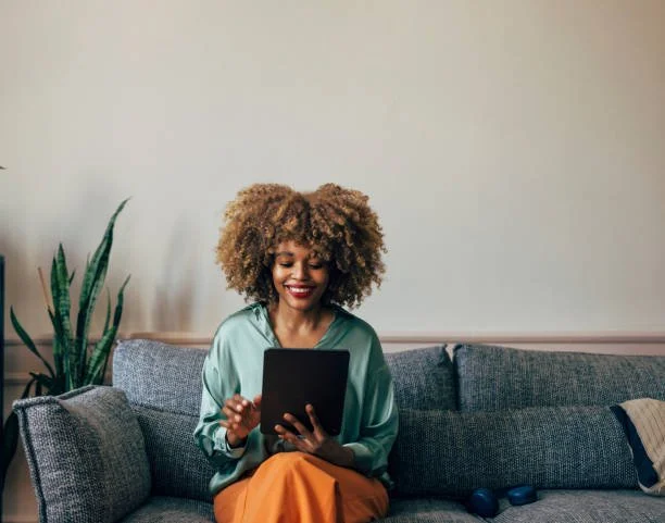 A woman with curly hair sitting on a gray couch, smiling as she looks at a tablet in her hands, with a potted plant to her left and a beige wall behind her.