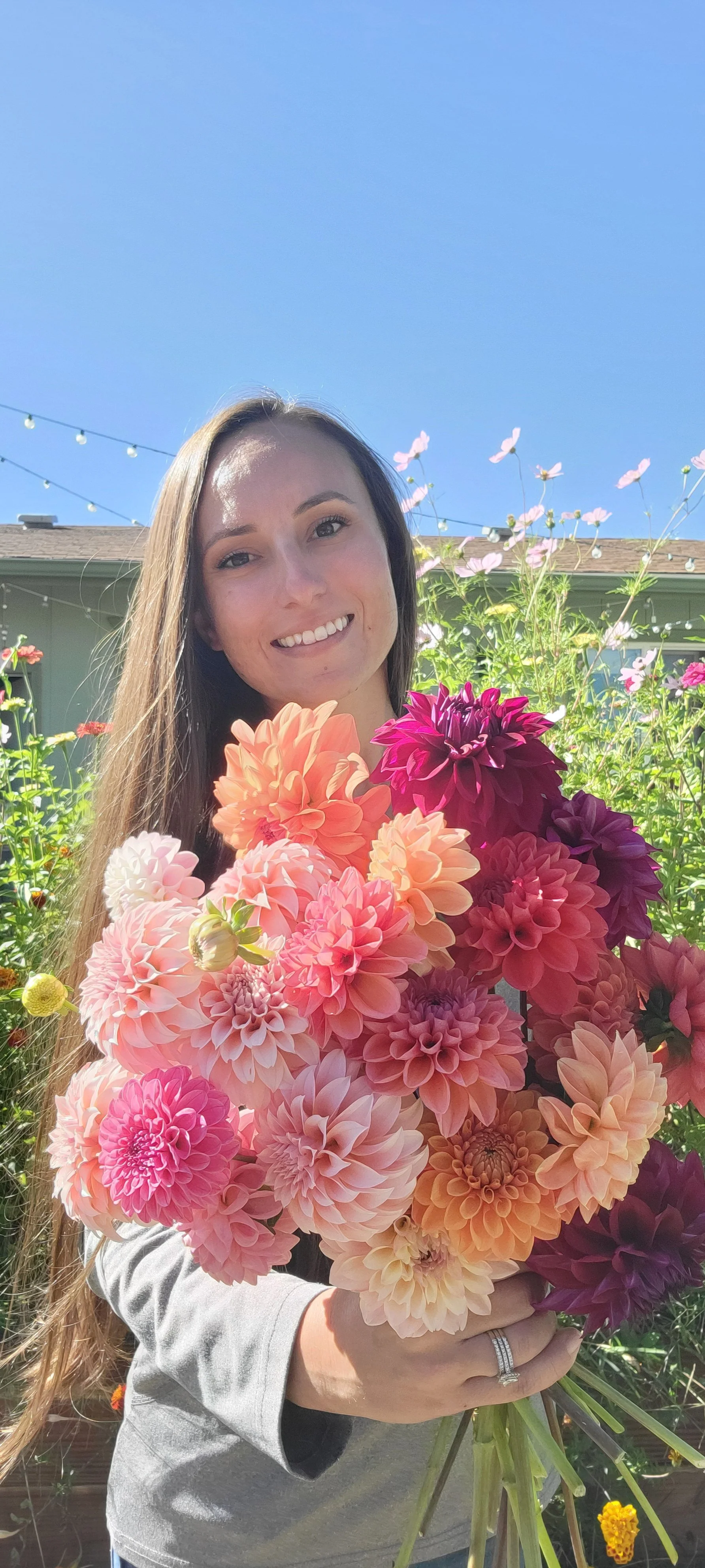 A woman smiling and holding a large bouquet of pink, orange, and purple dahlias outdoors on a sunny day.