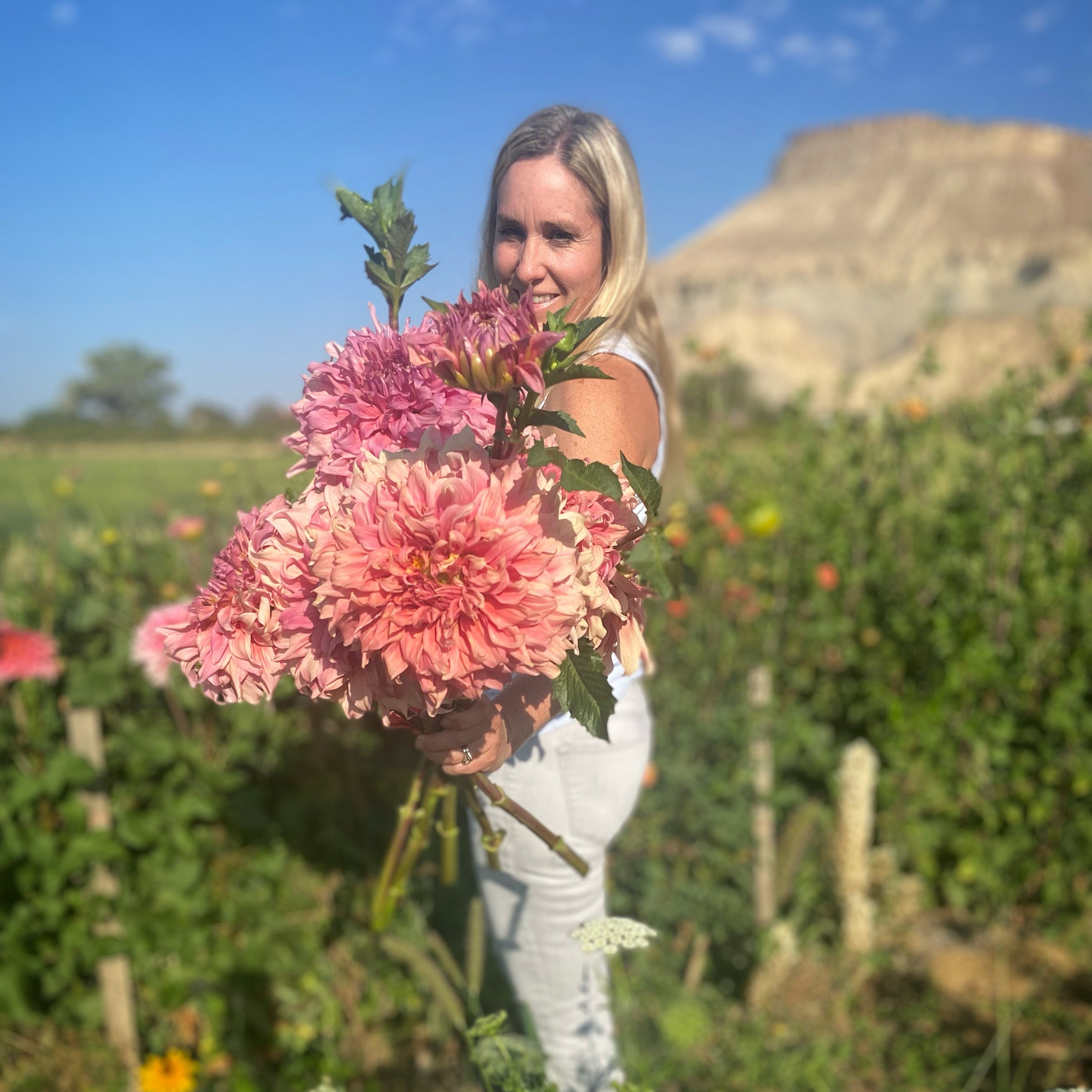 A woman with blonde hair smiles and holds a large bouquet of pink and lavender flowers in a garden with a mountain in the background.