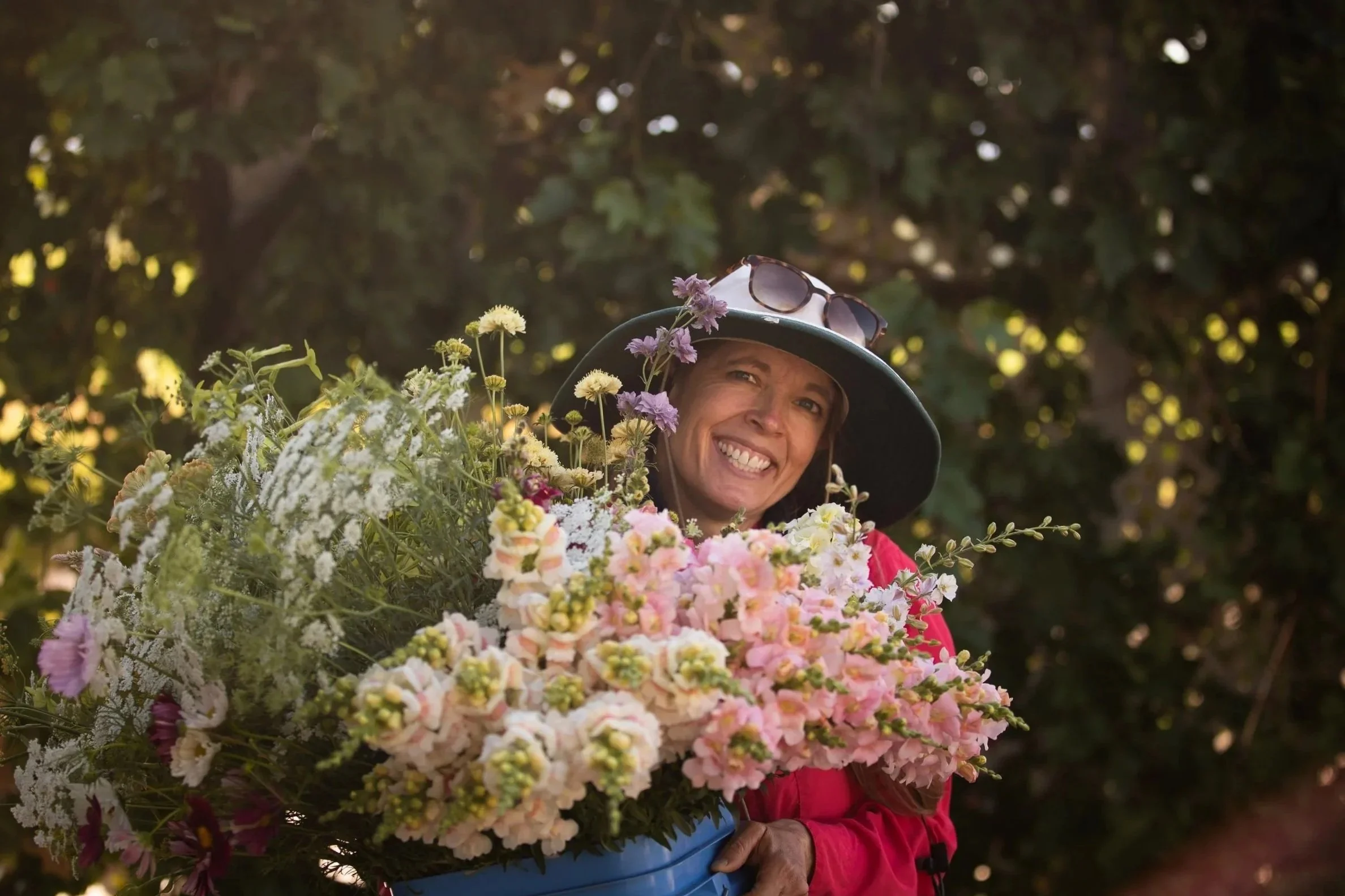 A woman wearing a wide-brimmed hat with sunglasses on top, smiling while holding a large bouquet of pink, white, and purple flowers against a background of dark green foliage.