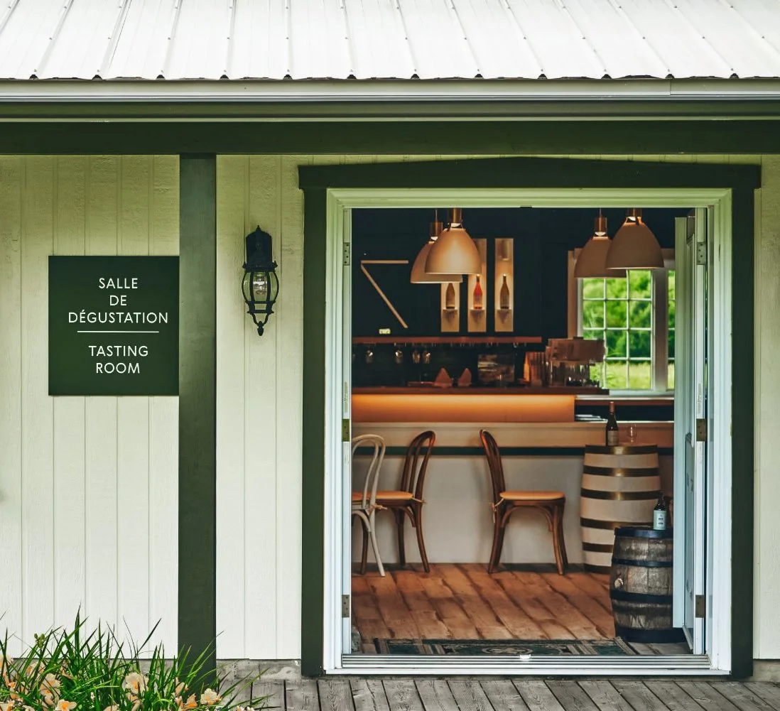 Salle de dégustation située dans une cabane en bois avec un signe en français et en anglais. Intérieur chaleureux en bois, chaises, étagères avec bouteilles, grande fenêtre avec vue sur la nature.