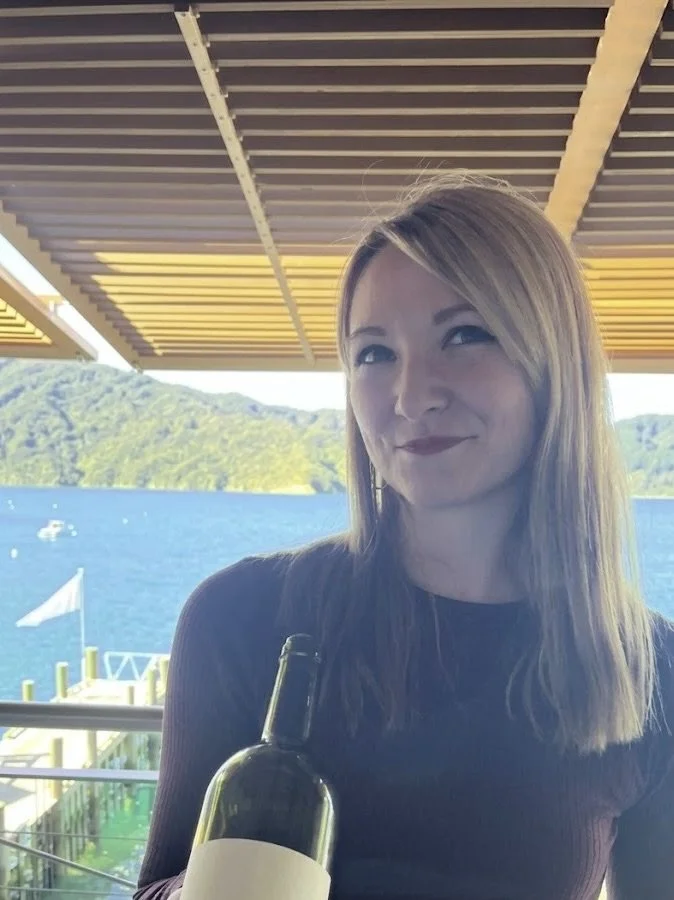 Une femme avec des cheveux longs, tenant une bouteille de vin, assise sous une structure en bois au bord de l'eau avec des montagnes en arrière-plan.