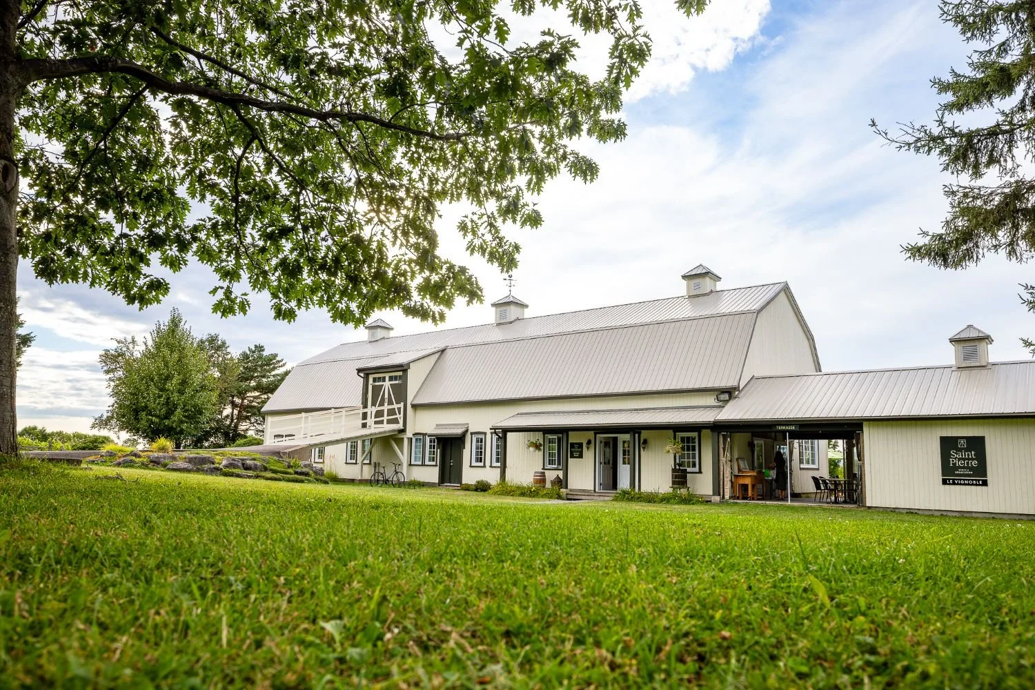Bâtiment principal du vignoble de style grange avec toit en métal, entouré d'arbres et de pelouse verte, sous un ciel partiellement nuageux, avec une terrasse où des personnes peuvent se retrouver.
