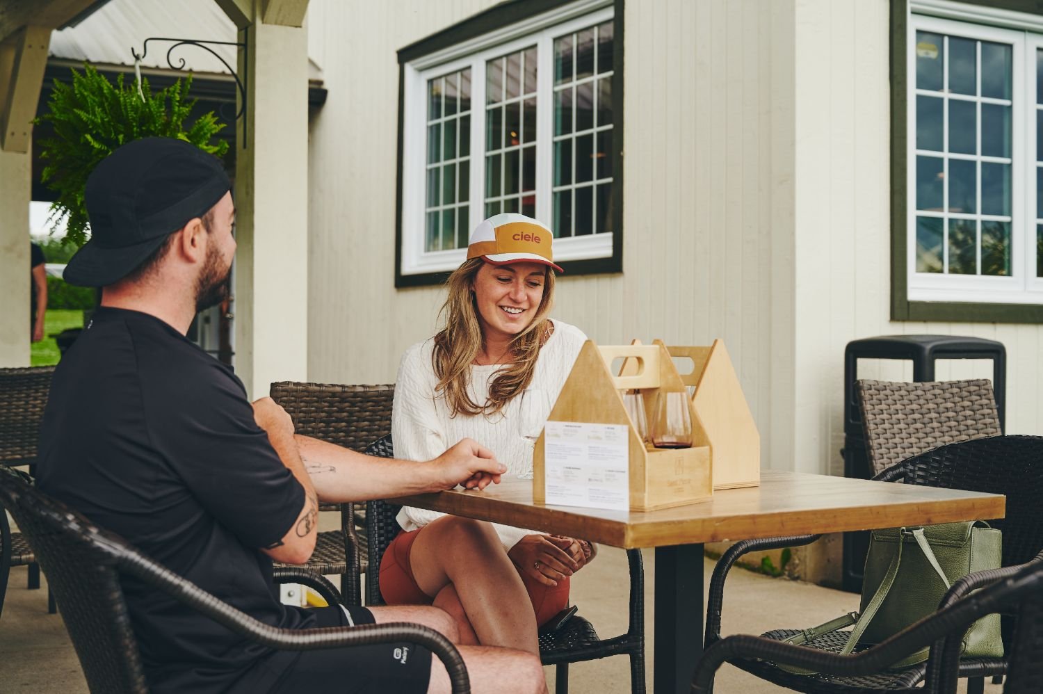 Deux personnes assises à une table sur la terrasse extérieure, discutant, avec une maison en arrière-plan. La femme porte un chapeau et sourit, l'homme est vêtu de noir et porte une casquette.