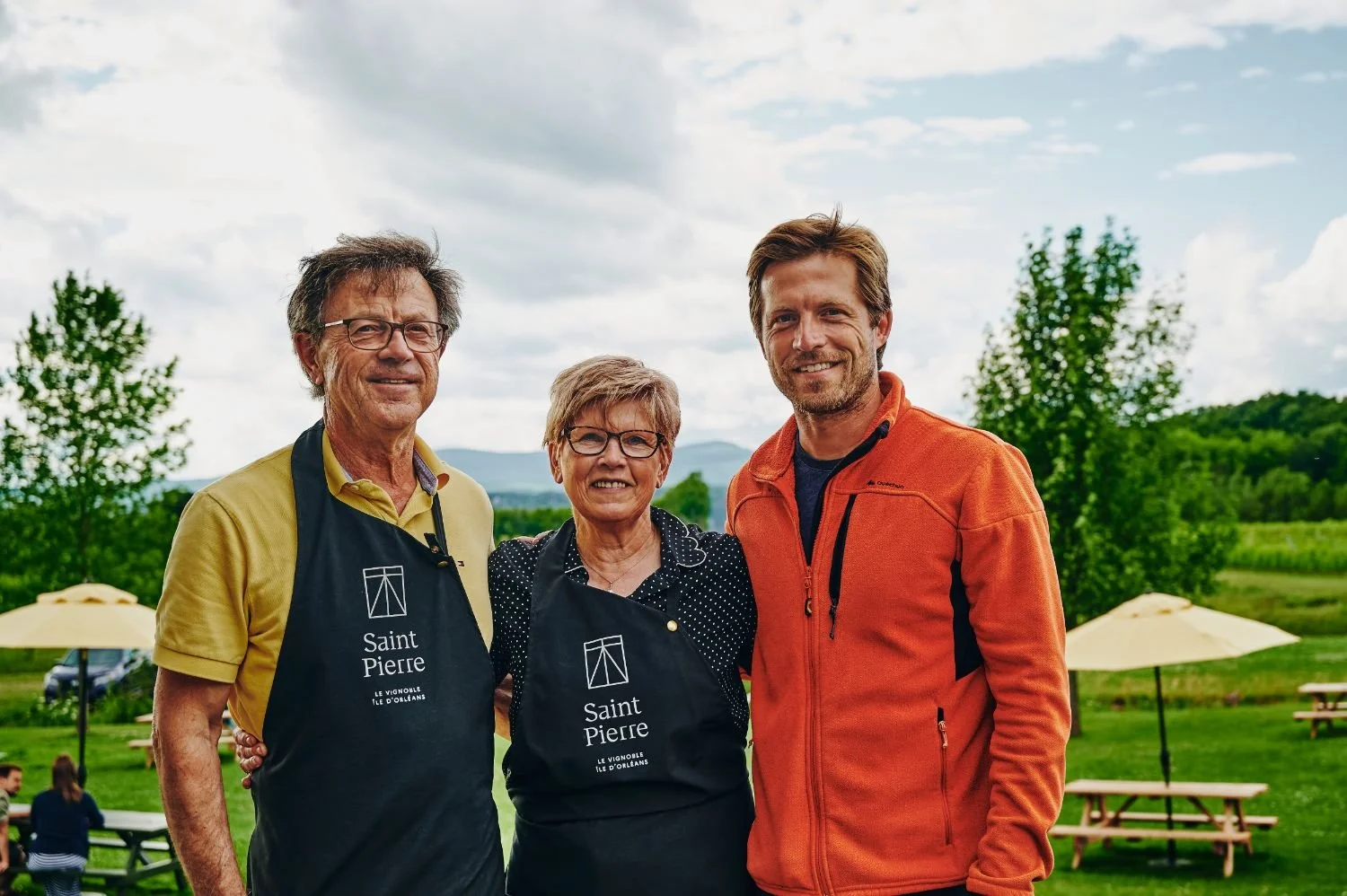 Trois personnes souriantes posent dans un jardin avec des parasols et des tables en bois, avec des arbres et des montagnes en arrière-plan.