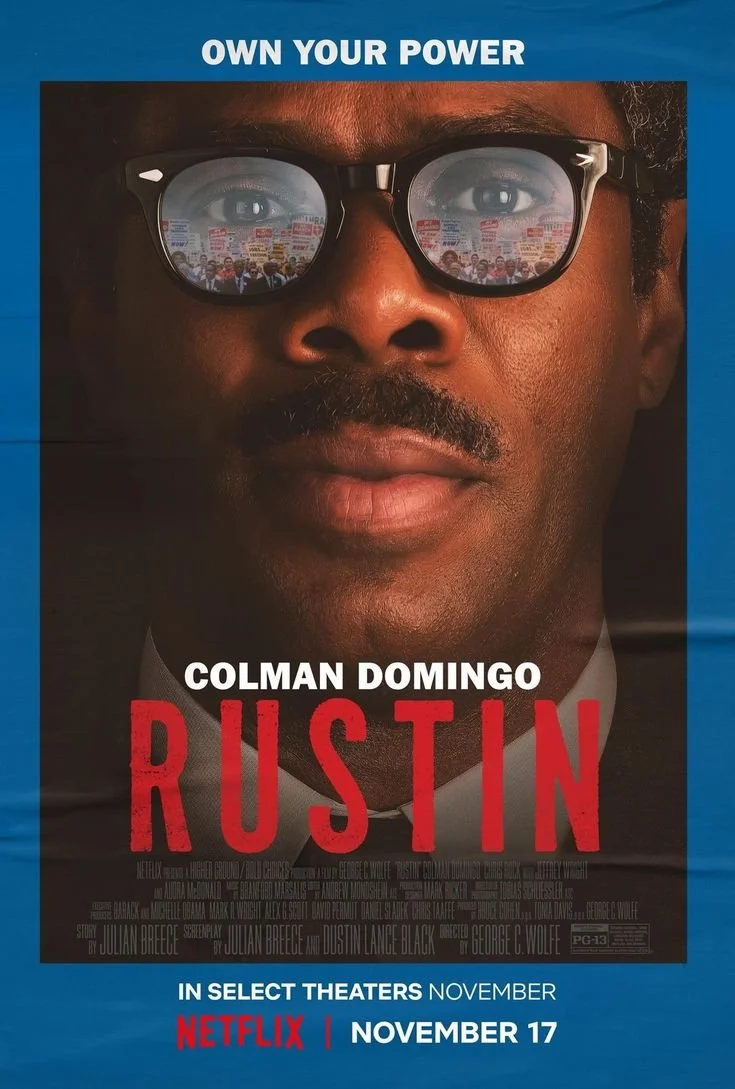 Close-up of a man's face with dark skin, wearing glasses that reflect a crowd holding signs, with text indicating "Own Your Power" and a promotion for the movie "Rustin" starring Colman Domingo, available in theaters and on Netflix starting November 17.
