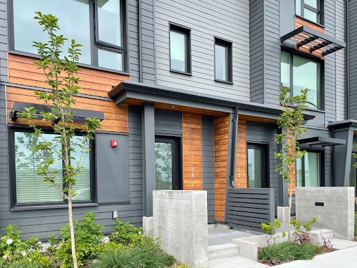 Modern townhouse with gray and wood exterior, small front garden with trees and shrubs, concrete steps leading to the door.