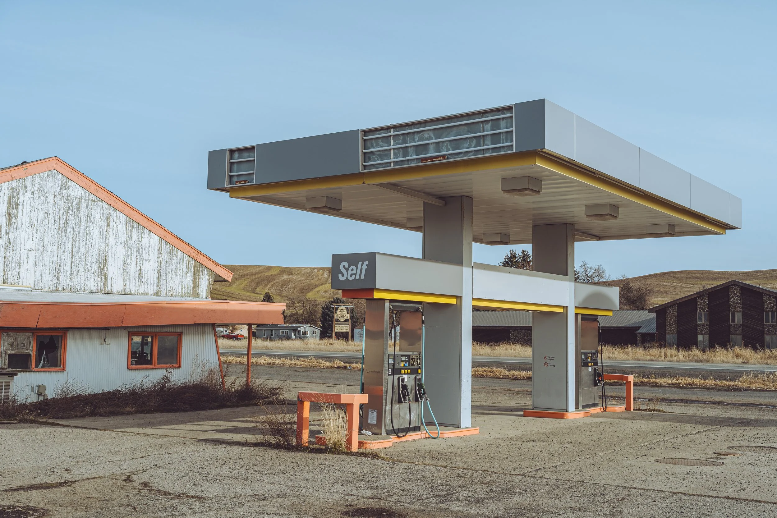 An old, empty gas station in a rural area under a clear blue sky with a few buildings and hills in the background.
