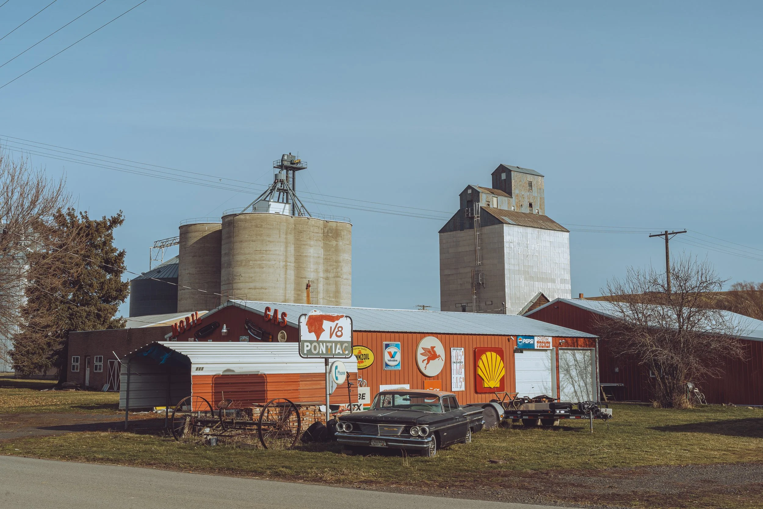 A rural roadside scene featuring an old barn with vintage signs including Shell and Pontiac, a classic black car, and large grain silos in the background under a clear blue sky.