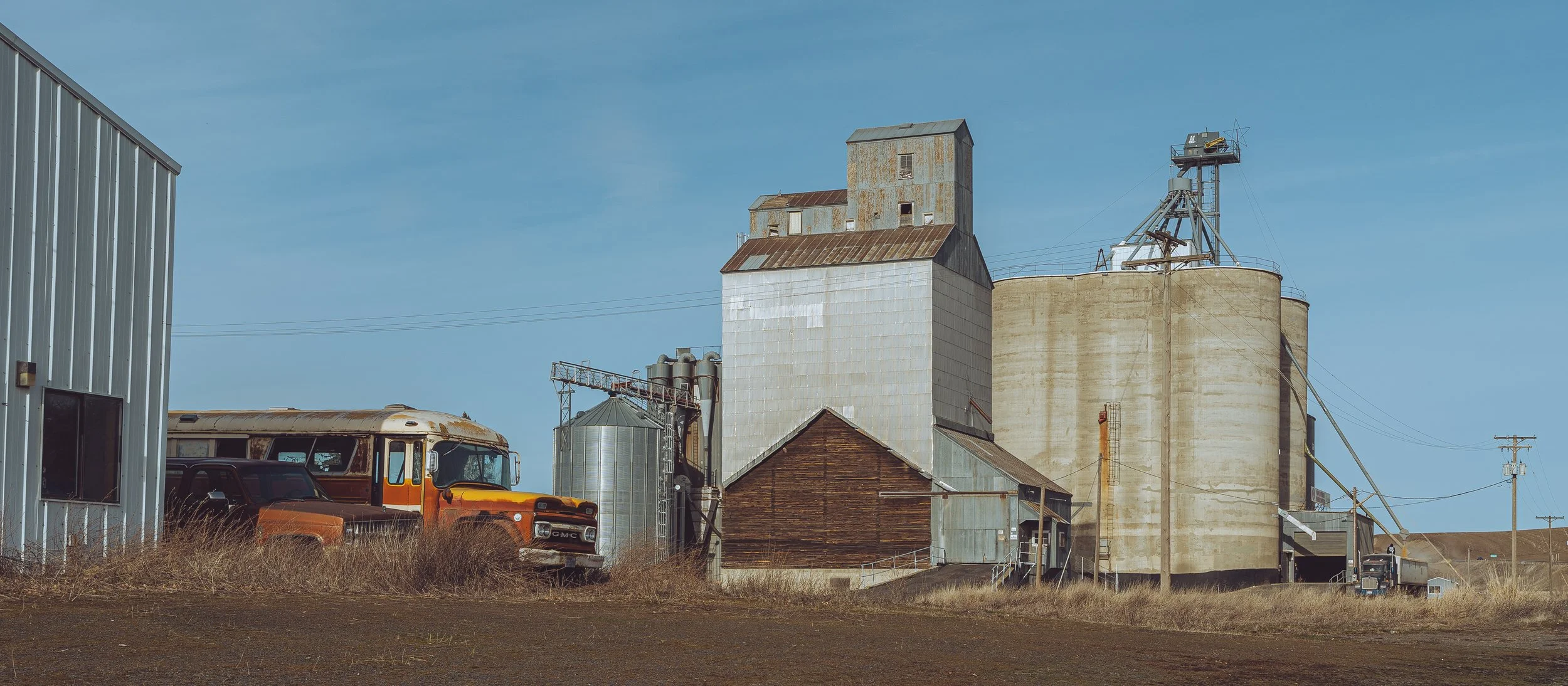 Old farm buildings, silos, and vintage vehicles outside on a rural landscape under a clear blue sky.