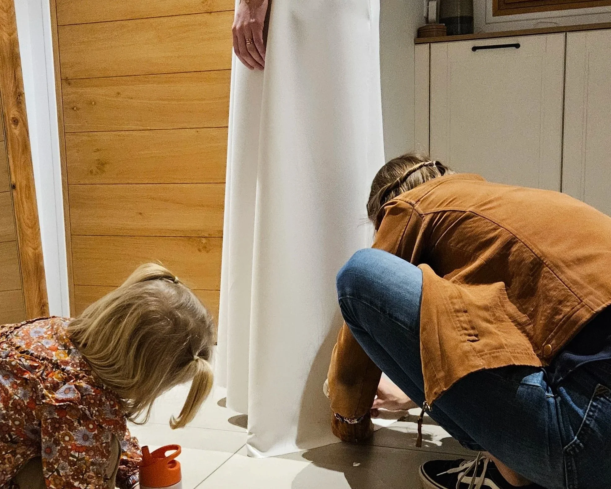 Une petite fille regarde Solenne, créatrice de robe de mairée sur mesure, travaillant au ras du sol, pour ajuster la longueur de la robe de mariée_Maison d'Héra