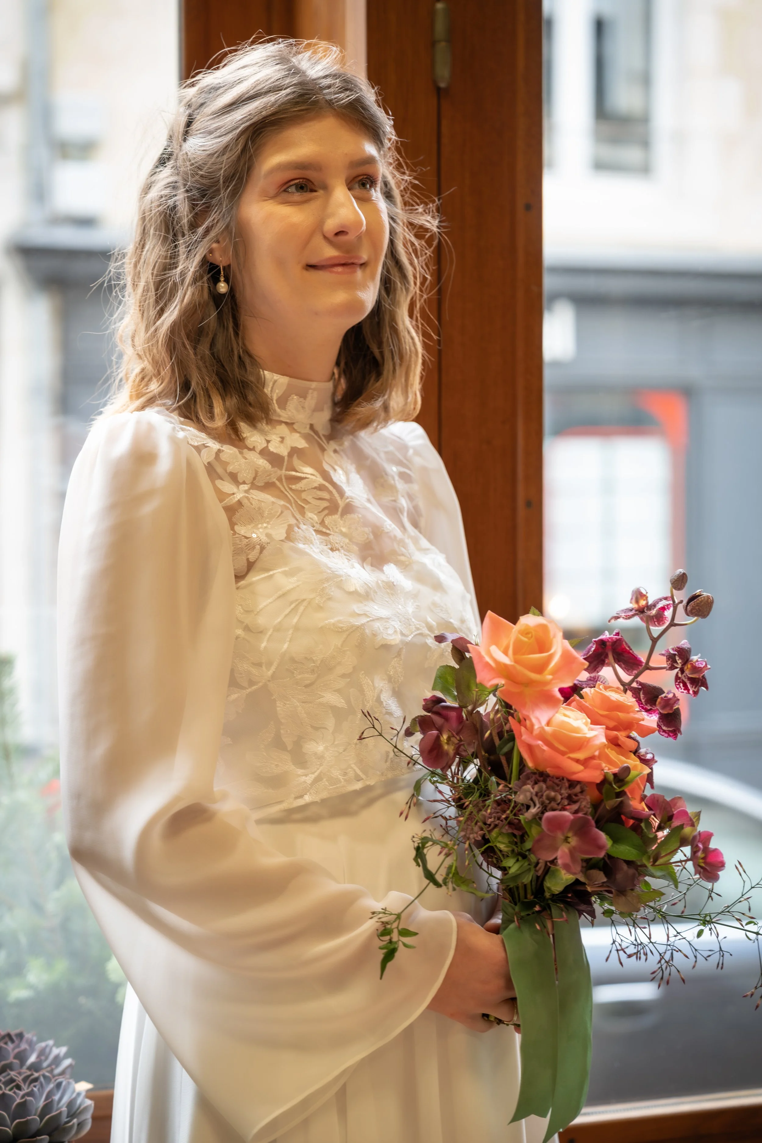 Une femme en robe de mariée blanche avec des détails en dentelle fleuraux, tenant un bouquet de fleurs orangées et violettes, devant une fenêtre en bois.