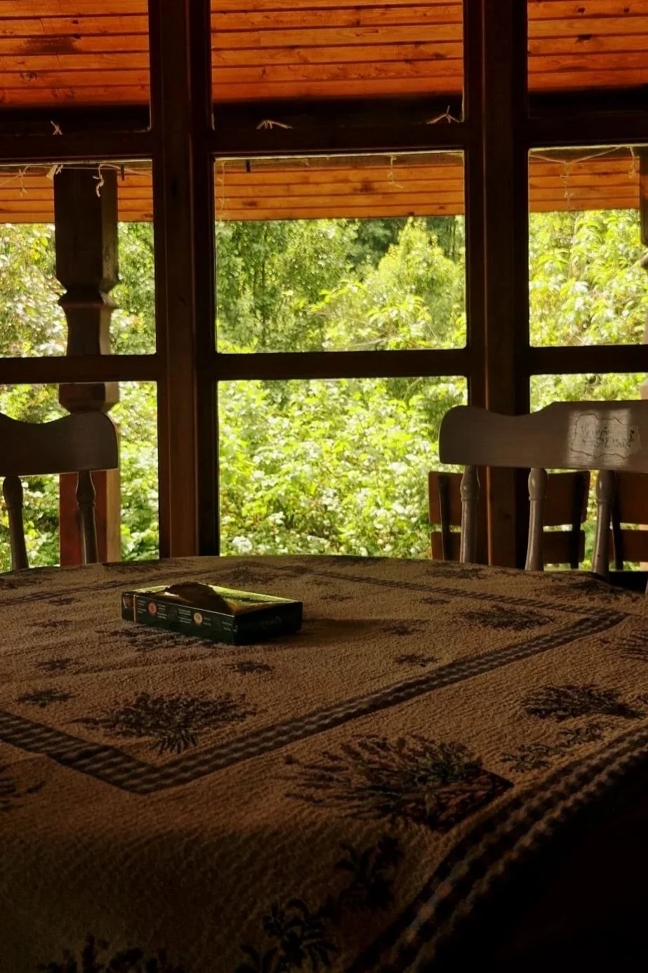A table with a patterned tablecloth near large windows overlooking lush green trees, with two wooden chairs and a small box or packet on the table.