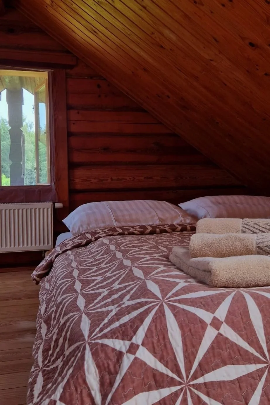 A cozy bedroom with a wooden paneled sloped ceiling and wooden walls, a window with a view of greenery outside, a radiator beneath the window, a bed with a patterned bedspread and pillow, and neatly folded towels on the bed.
