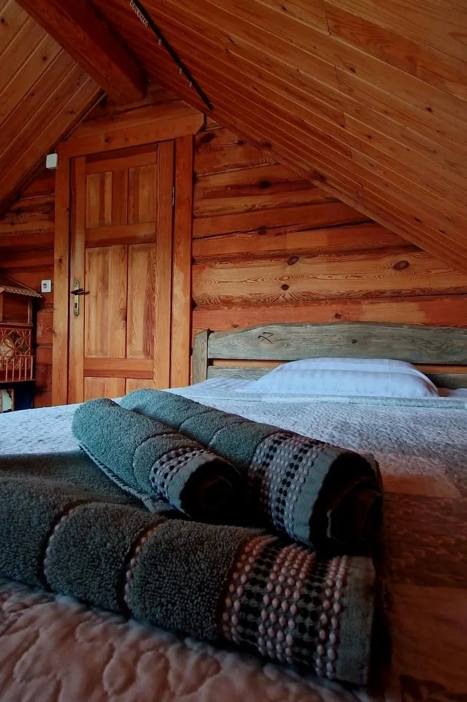 A cozy bedroom with wooden panel walls and sloped ceiling, featuring a bed with white bedding and rolled towels in the foreground.