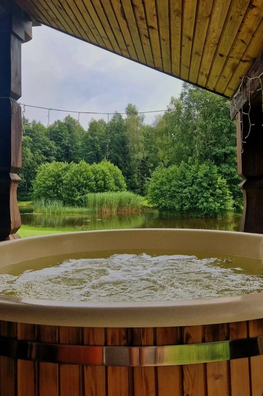 A hot tub with bubbling water on a wooden porch overlooking a lush green landscape with trees and a pond.