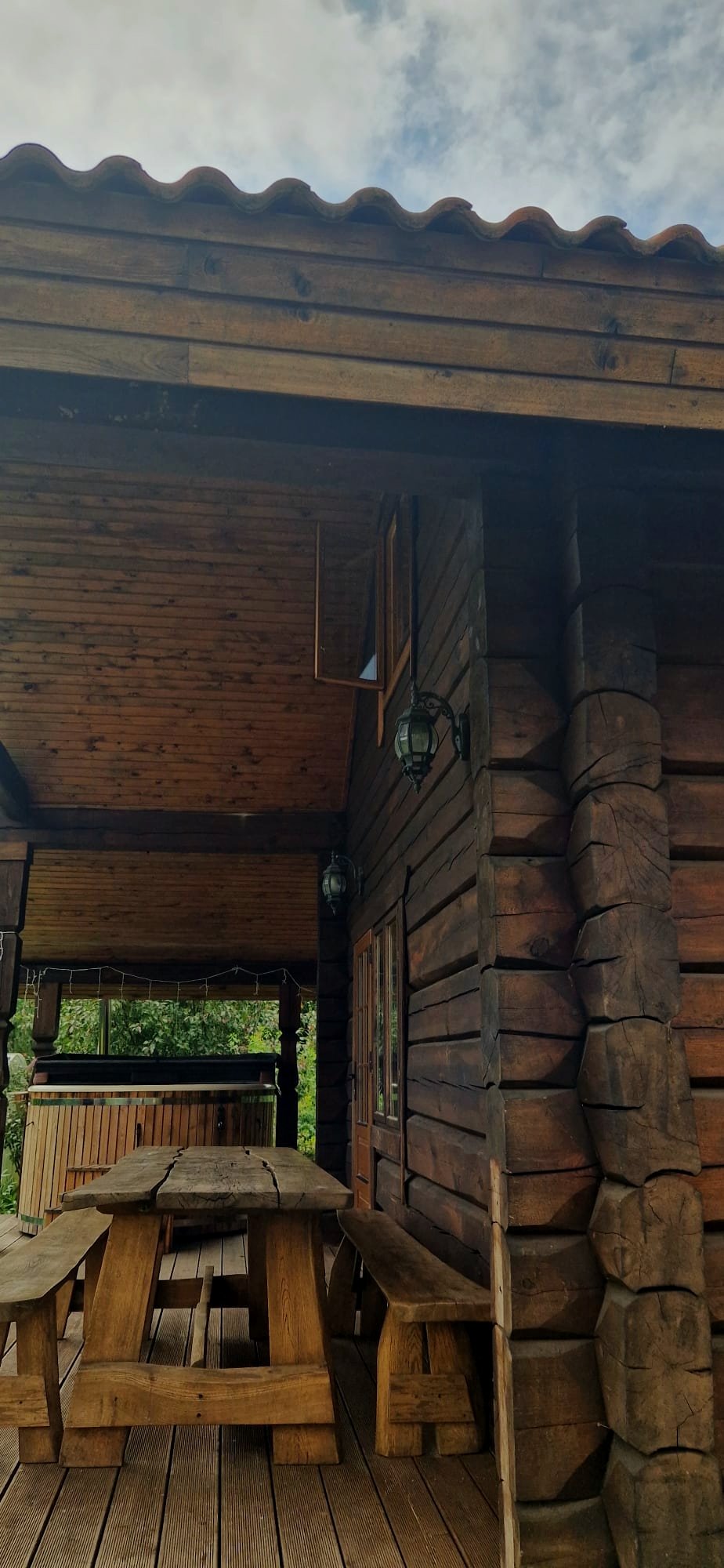 Wooden outdoor patio with a picnic table, benches, and a hot tub in a rustic cabin setting, with trees in the background and an overcast sky.