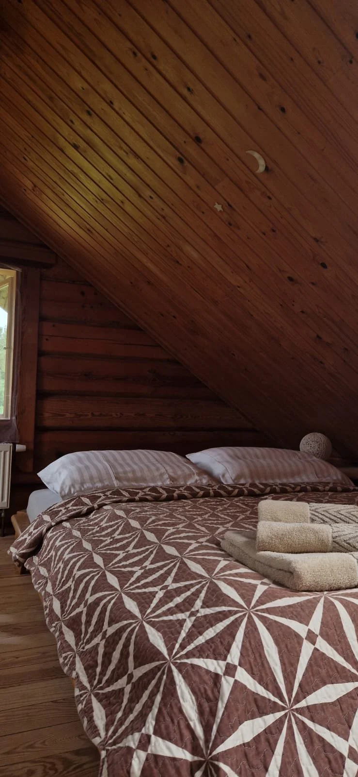 A cozy bedroom with a wooden sloped ceiling, a bed with patterned brown and white bedding, and towels placed on the bed.
