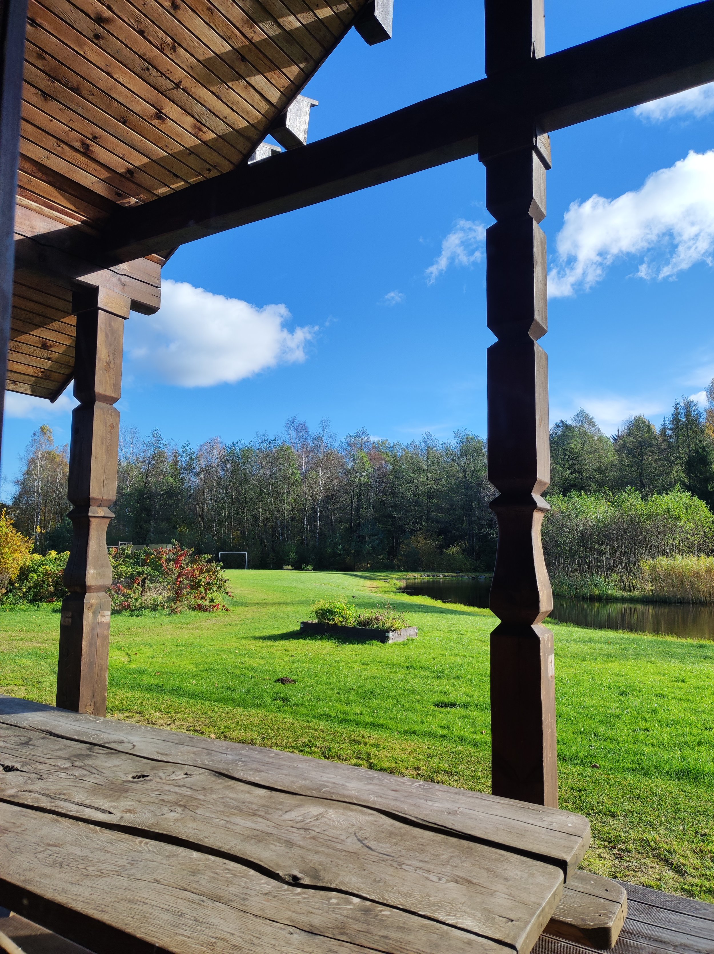 A view from a wooden porch overlooking a green field, trees, a pond, and a blue sky with some clouds.