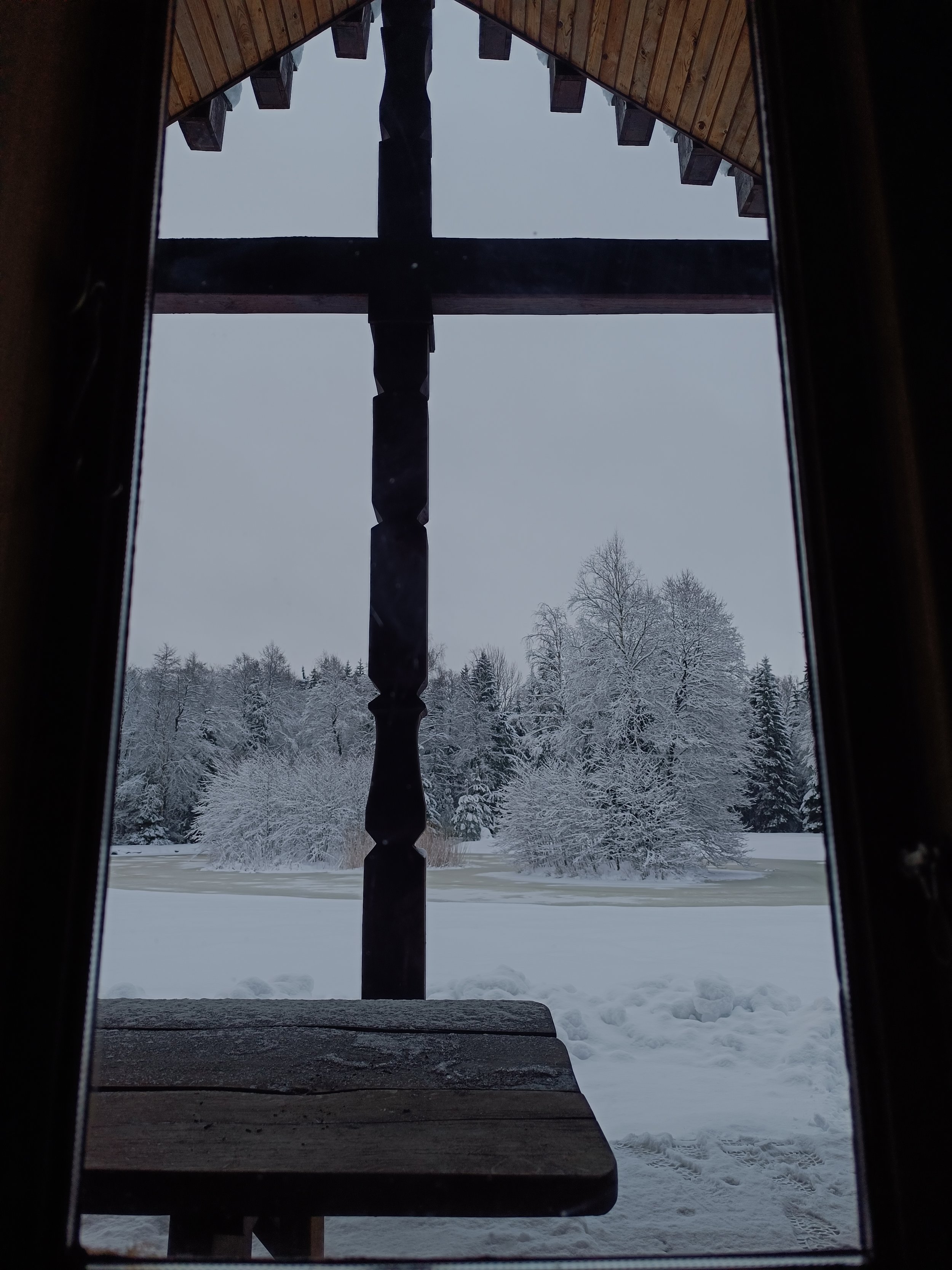 View of snowy landscape with trees covered in snow viewed from inside a wooden cabin, framed by the window with a wooden bench below.