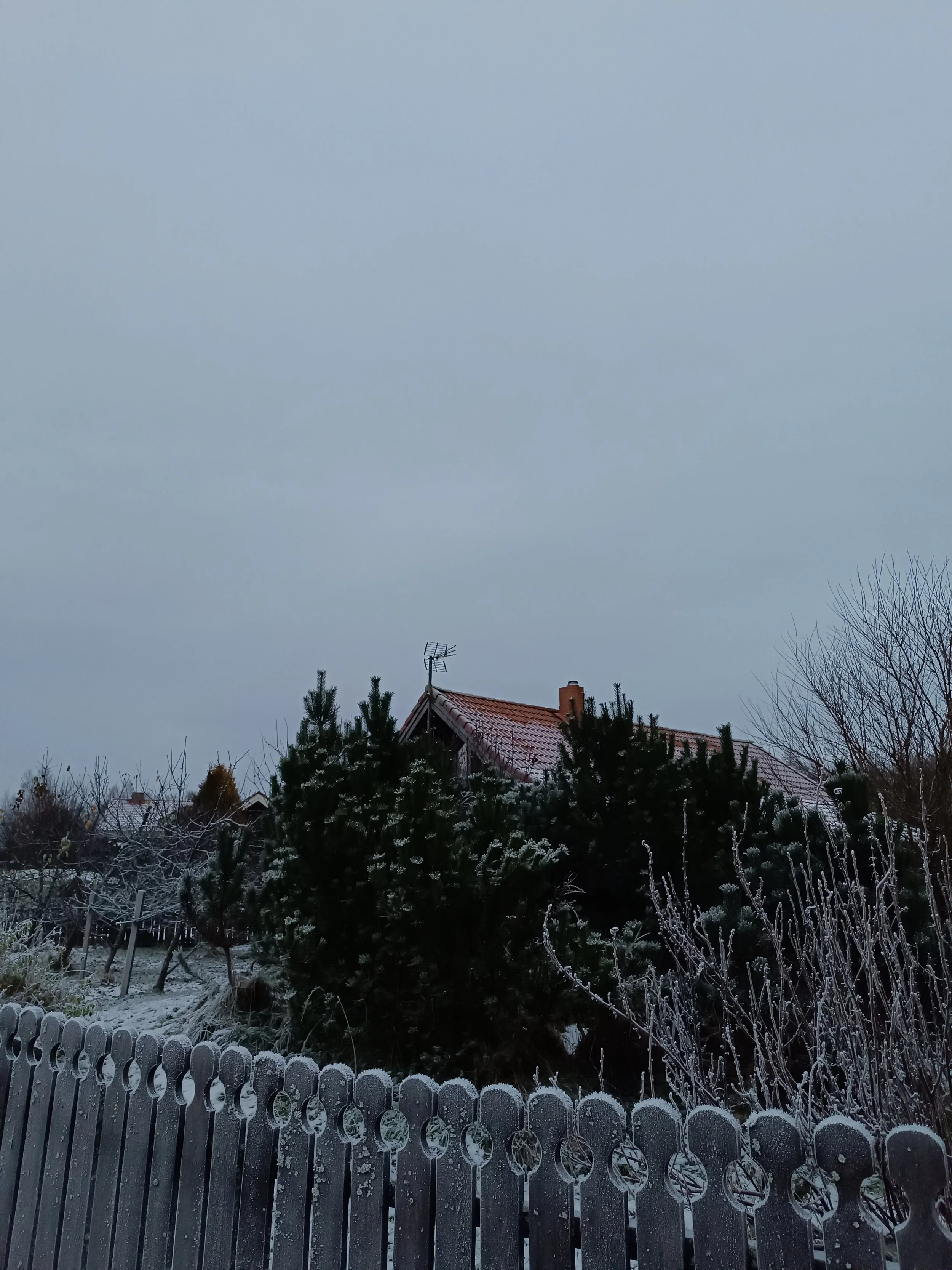 A winter scene with a house, snow-covered trees, and a wooden fence in the foreground. The house has a red-tiled roof and an outdoor antenna.