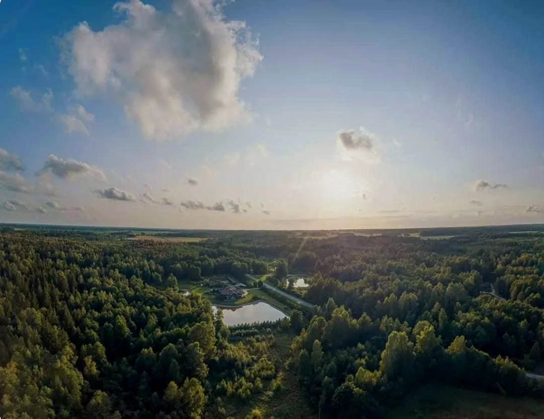 Aerial view of a lush green landscape with dense forest, two lakes, and a small cluster of buildings, under a partly cloudy sky at sunset.