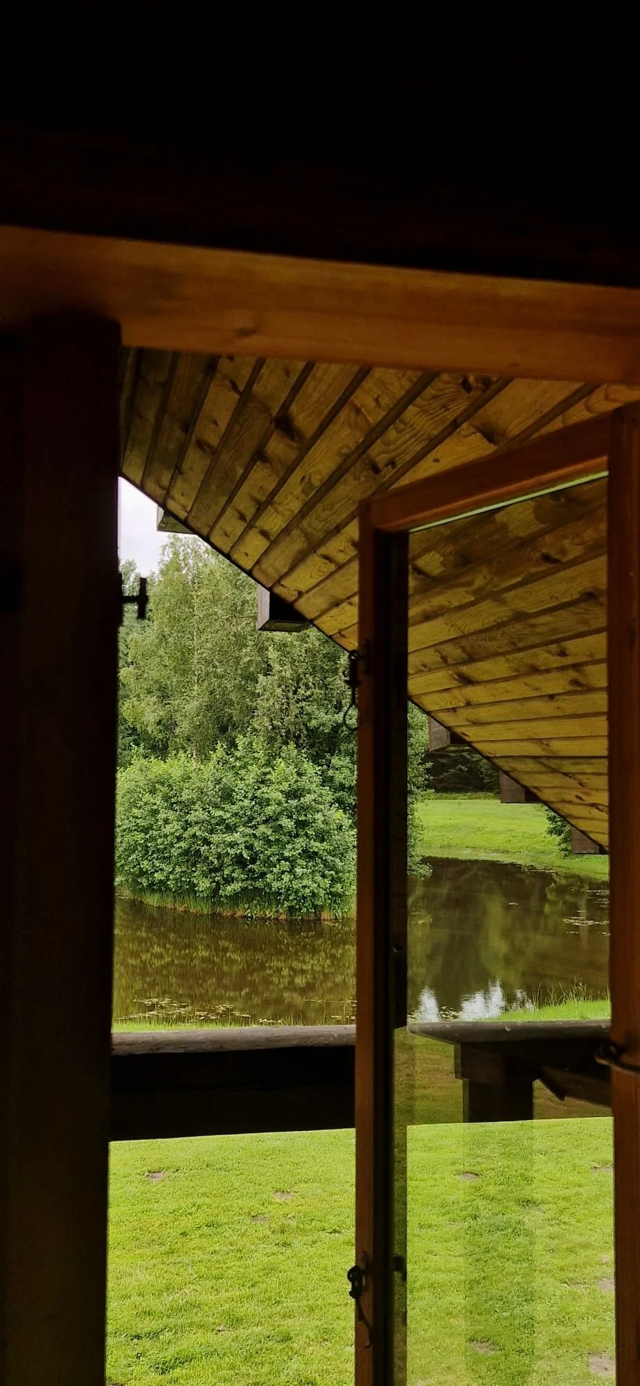 View of a river through an open door of a wooden house with a sloped roof, green grass, trees, and cloudy sky in the background.