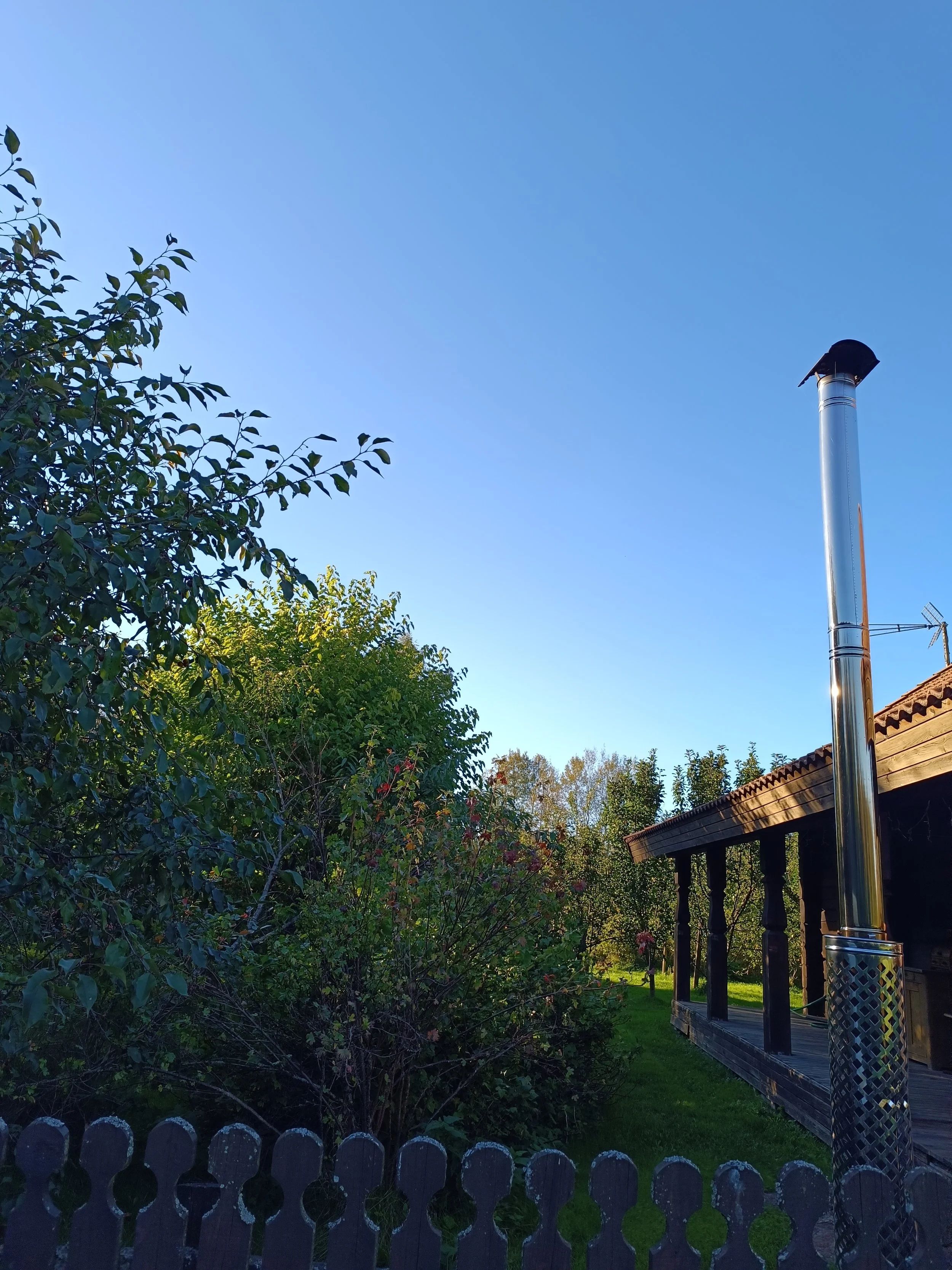 View of a garden with trees and bushes, a wooden fence in the foreground, and a house with a porch on the right. A metallic chimney with a cap stands prominently to the right. Clear blue sky above.