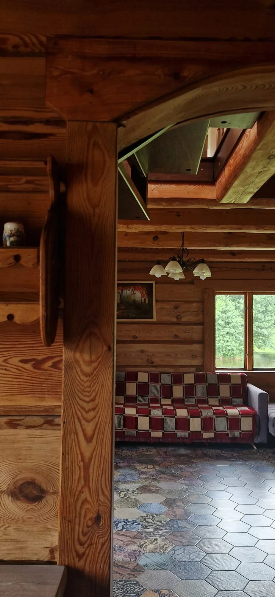 Interior view of a rustic wooden cabin with a sofa, a window showing greenery outside, a hanging chandelier, and a patterned tile floor.
