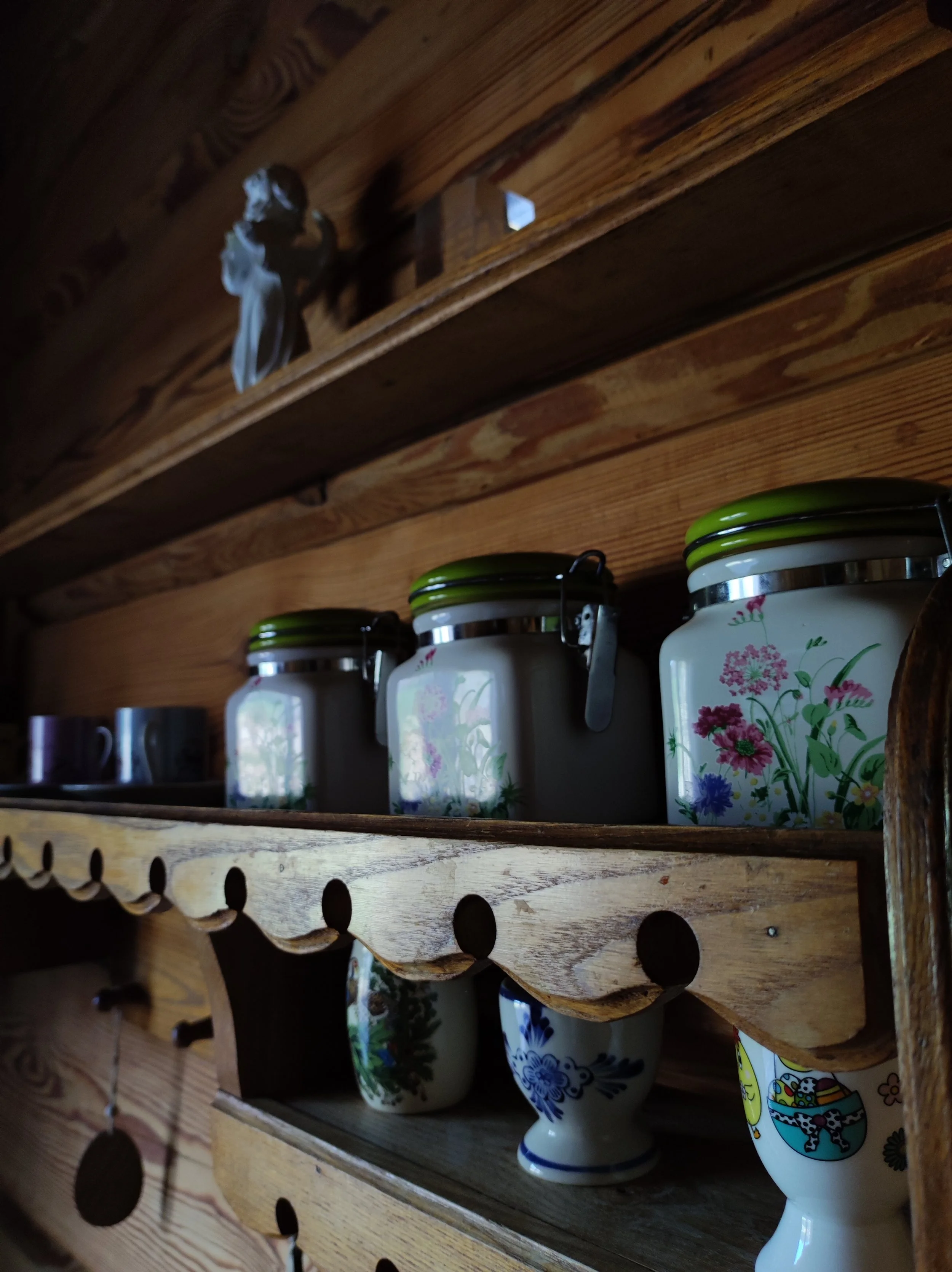 Wooden kitchen shelf with floral ceramic jars on top and painted ceramic cups on the bottom shelf, wooden wall in the background.