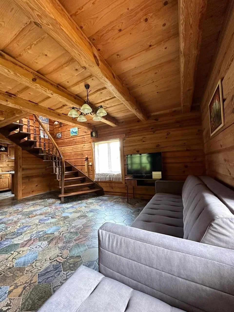 A cozy living room with wood-paneled walls and ceiling, a multi-colored hexagonal tile floor, a gray sofa, a small TV on a stand, a window with a curtain, a ceiling light fixture, and a wooden staircase leading to an upper level.