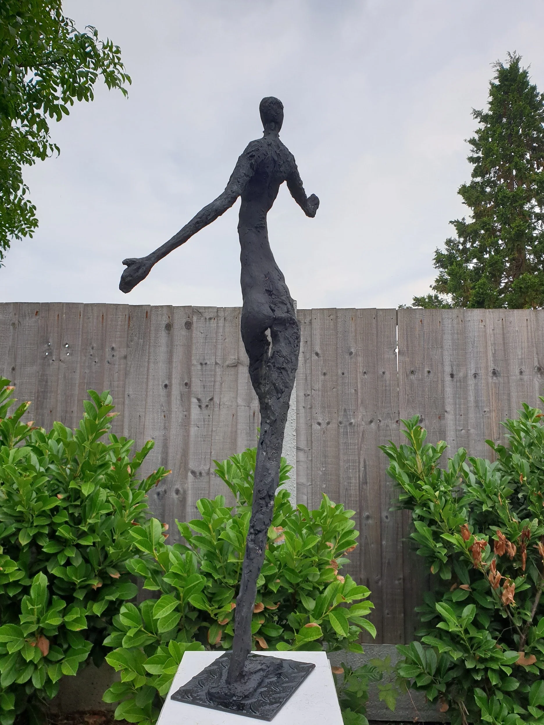 A tall, abstract black sculpture of a woman running or dancing, placed outdoors in front of a wooden fence and greenery.