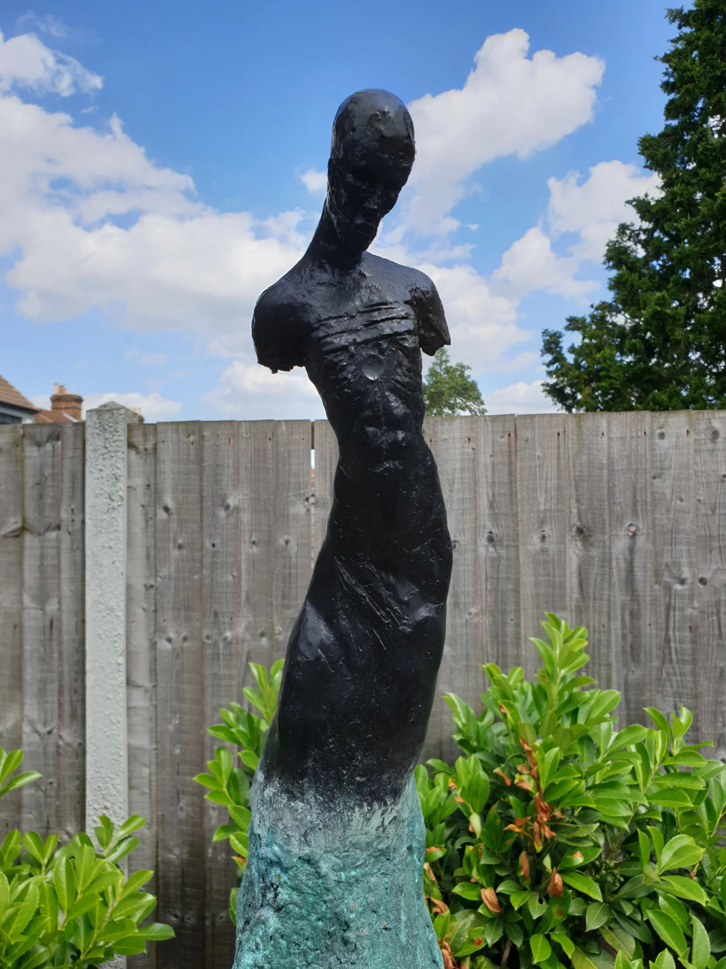 Black abstract sculpture of a human figure with an elongated head, standing outdoors in front of a wooden fence, with green bushes and trees, blue sky, and clouds in the background.