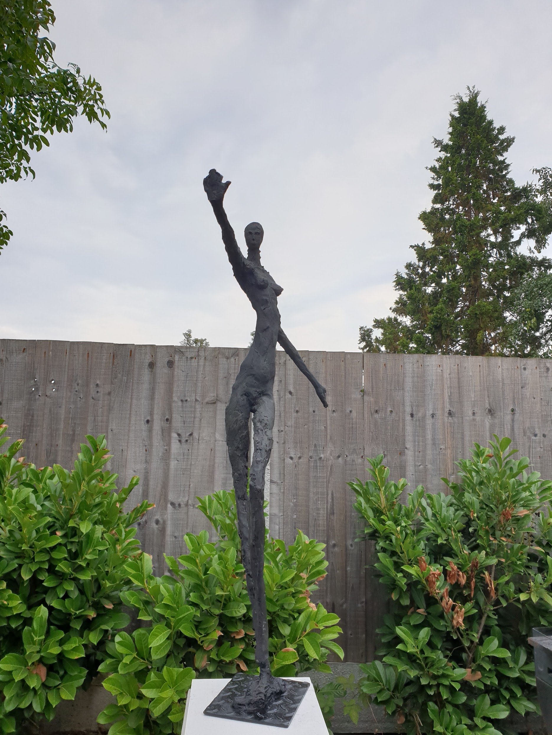 A modern abstract sculpture of a tall, slender woman with one arm raised, standing outdoors against a wooden fence and trees, with shrubs at the base.