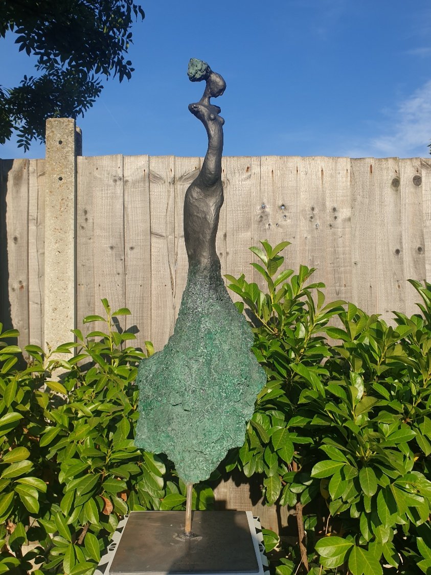 A metal sculpture resembling a tall, thin, abstract giraffe with a small head, situated outdoors against a wooden fence and surrounded by green bushes, with a blue sky overhead.