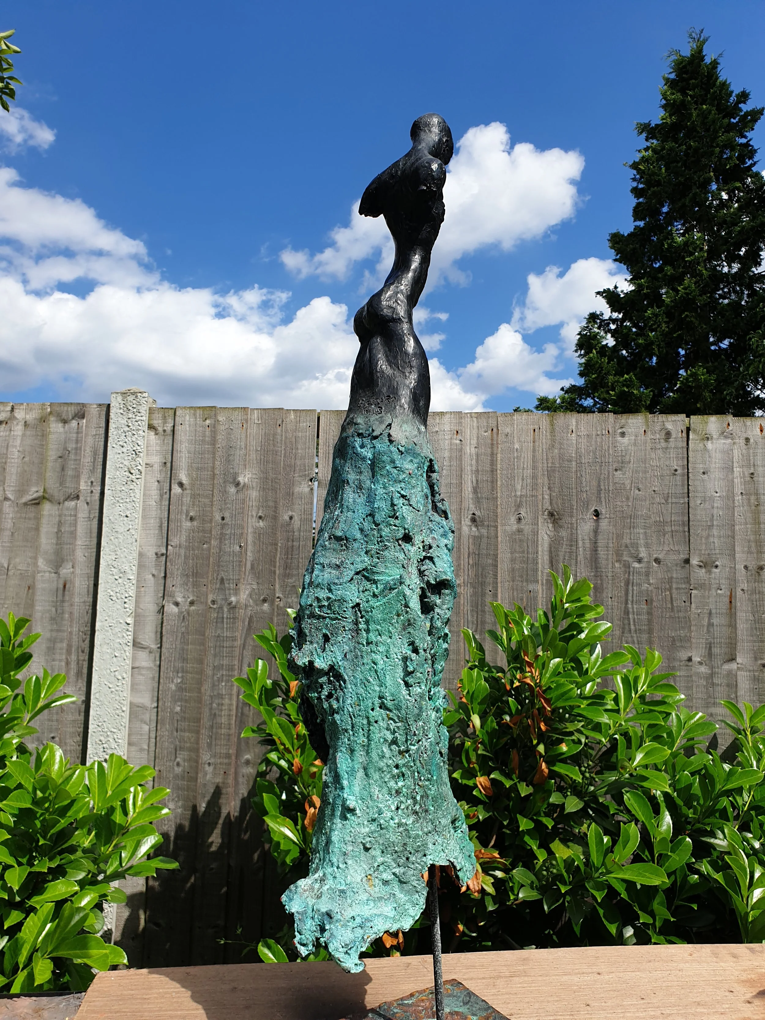 Outdoor sculpture of a tall, abstract female figure with a metallic finish, set against a wooden fence, green bushes, and a partly cloudy sky.