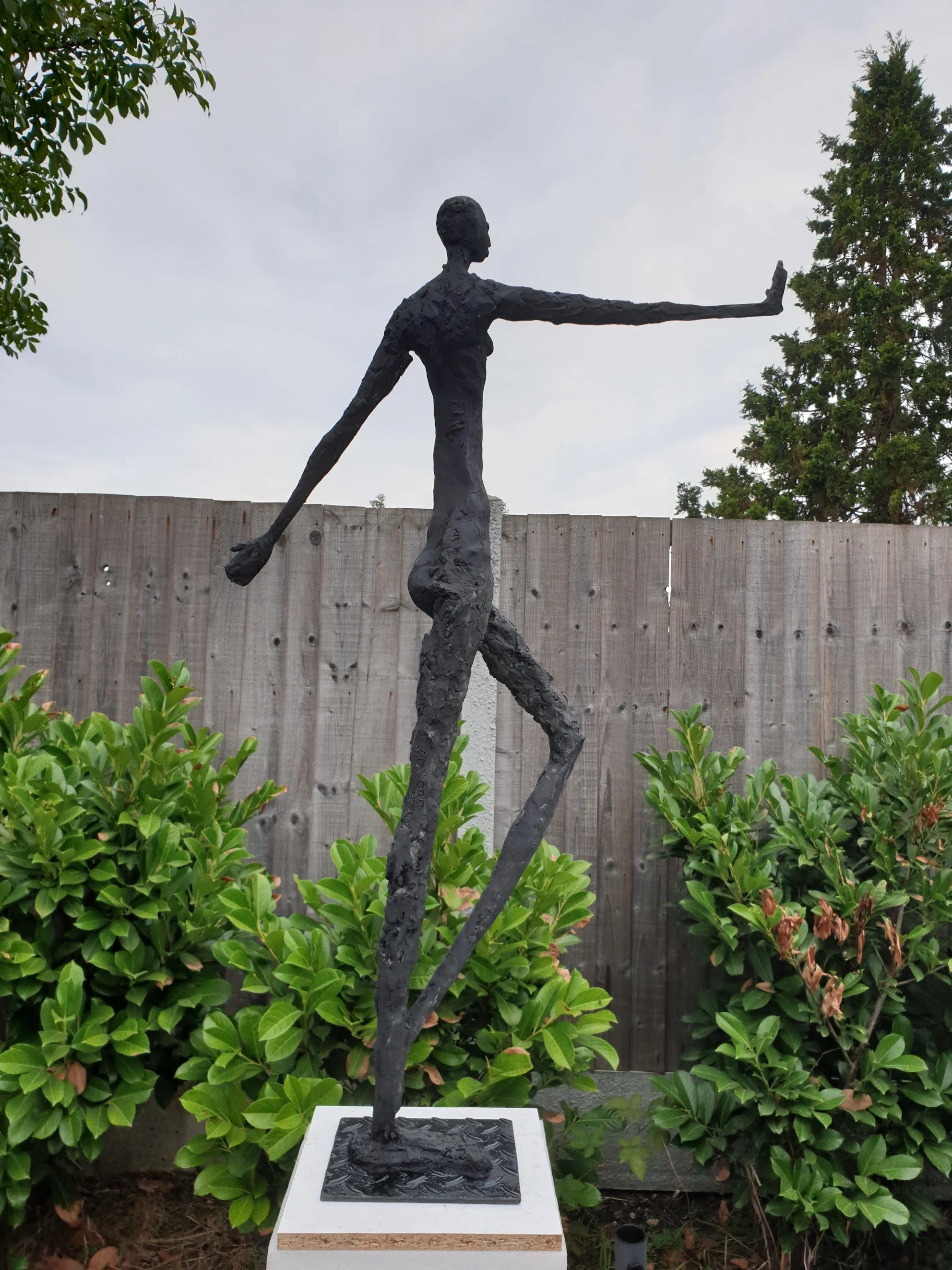 A tall bronze sculpture of a slender woman with extended arms, standing on one leg, with the other leg and arm stretched out, outdoors near greenery and a wooden fence.