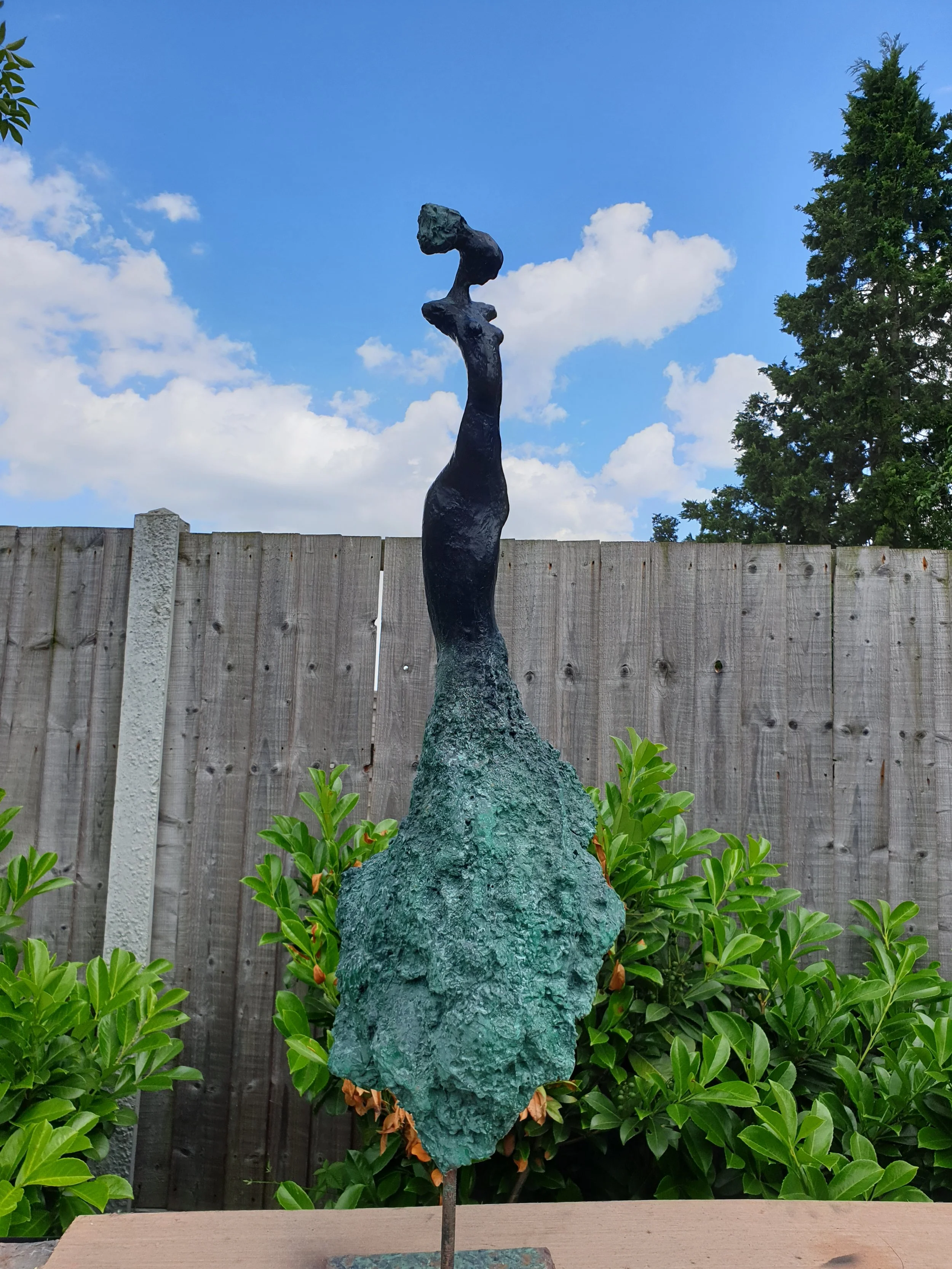 Abstract outdoor sculpture of a tall, elongated figure with a small head, dark and textured, set against a partly cloudy sky and surrounded by green bushes and a wooden fence.
