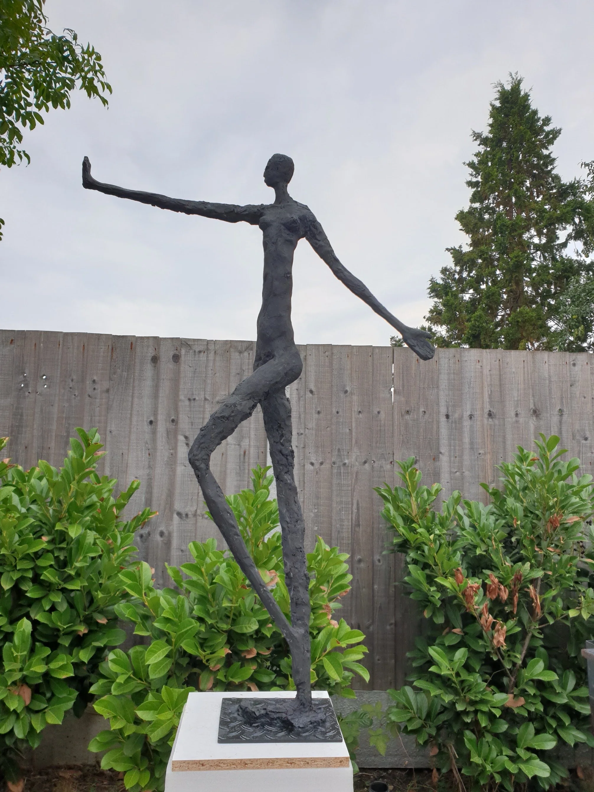 A tall abstract sculpture of a human figure with arms outstretched, standing on a pedestal in an outdoor garden, with a wooden fence and trees in the background.