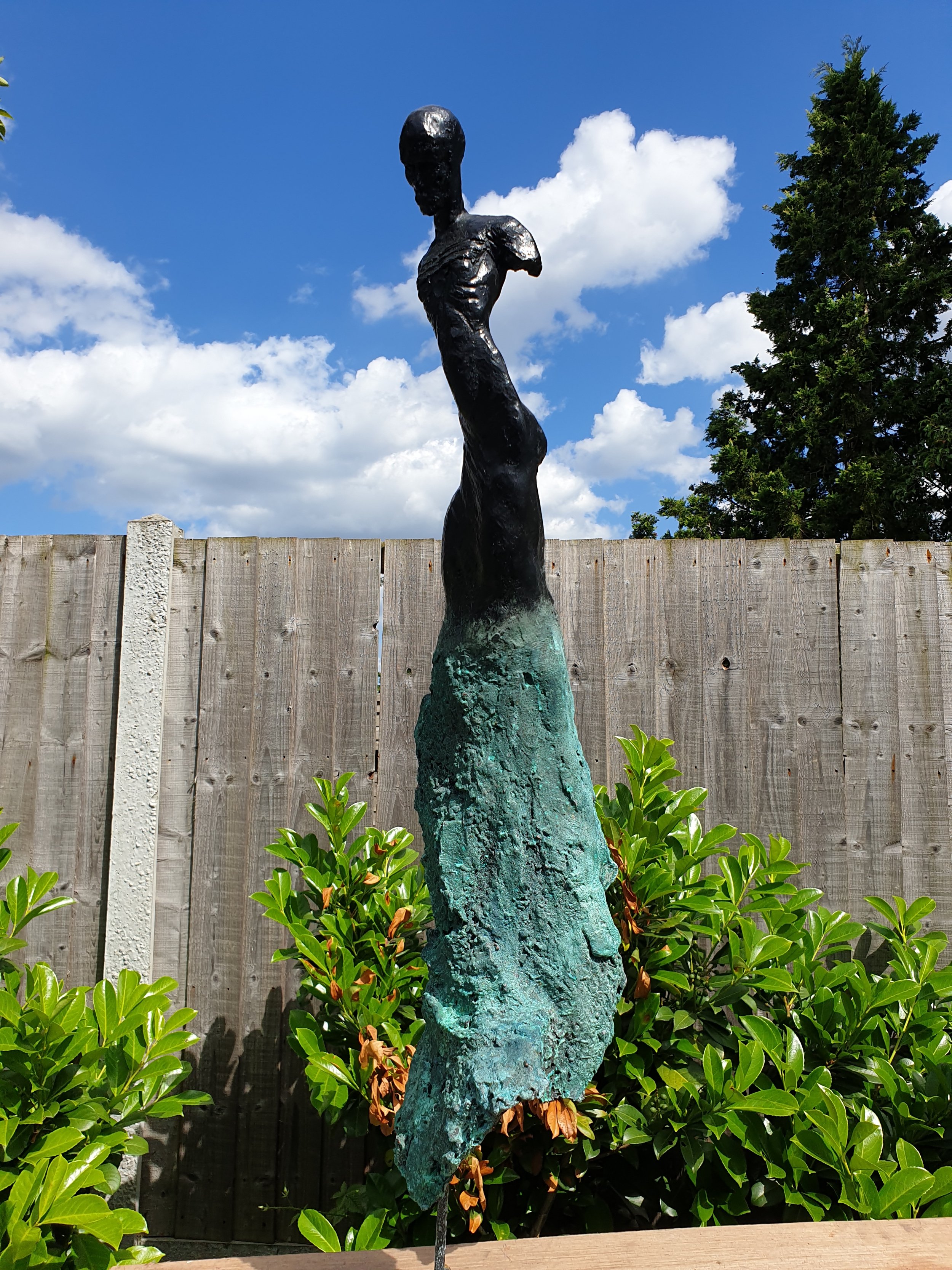 Outdoor sculpture of a slender woman on a tall, textured base, set against a blue sky with white clouds and a wooden fence, surrounded by green bushes.