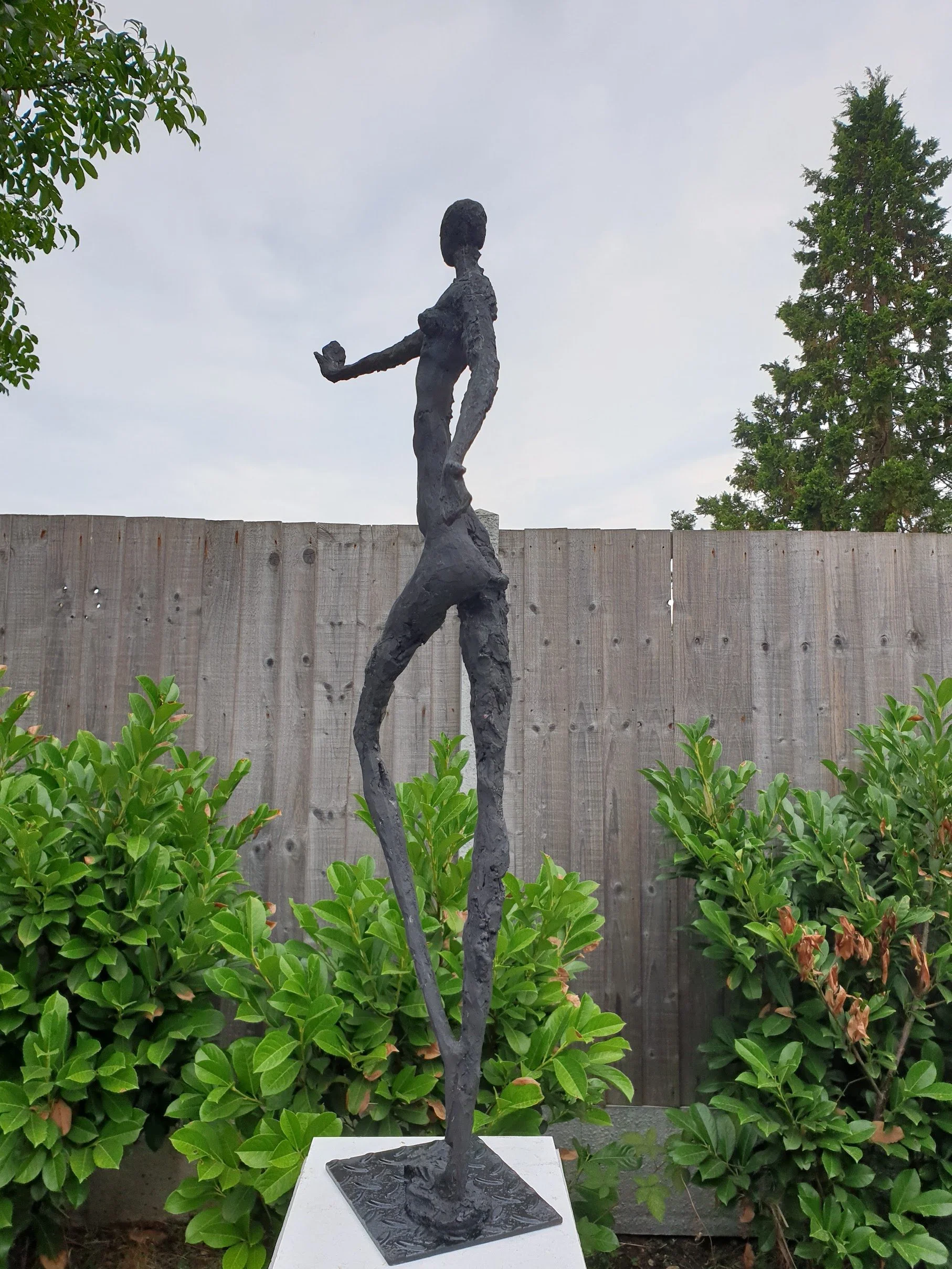 A tall, abstract sculpture of a human figure with one arm extended, set outdoors against a wooden fence and surrounded by greenery.